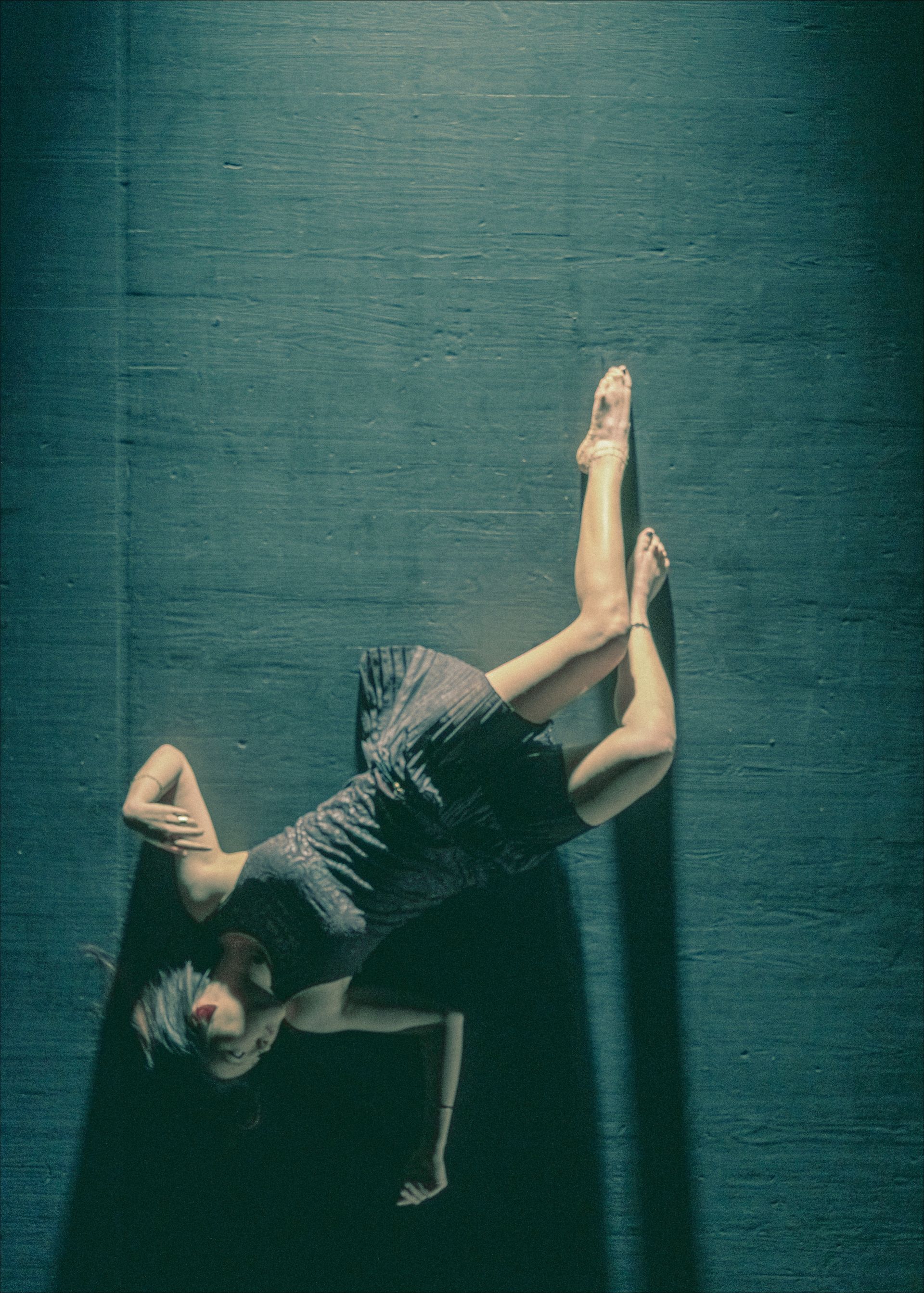 Woman in dark dress on a dark floor, in a dance pose with legs extended upward and arms crossed.