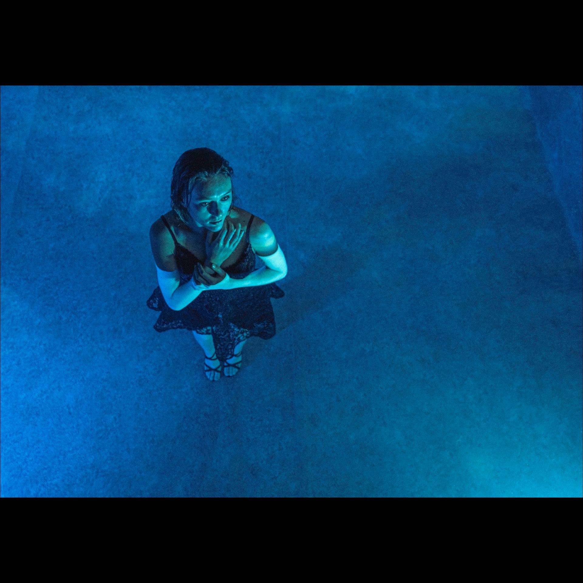 Woman in dark dress with hands clasped, lit in blue light, standing in water.