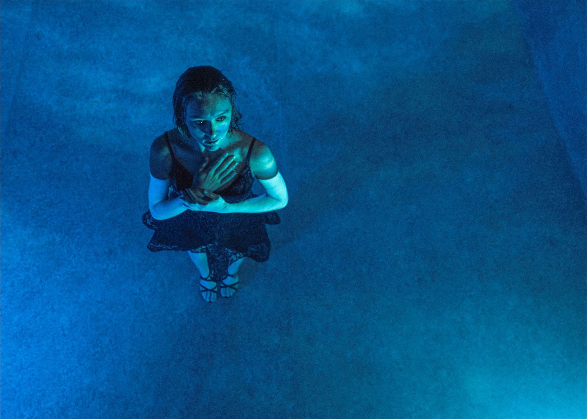 Woman in a dark dress, arms crossed, lit by blue light, standing in a hazy atmosphere.