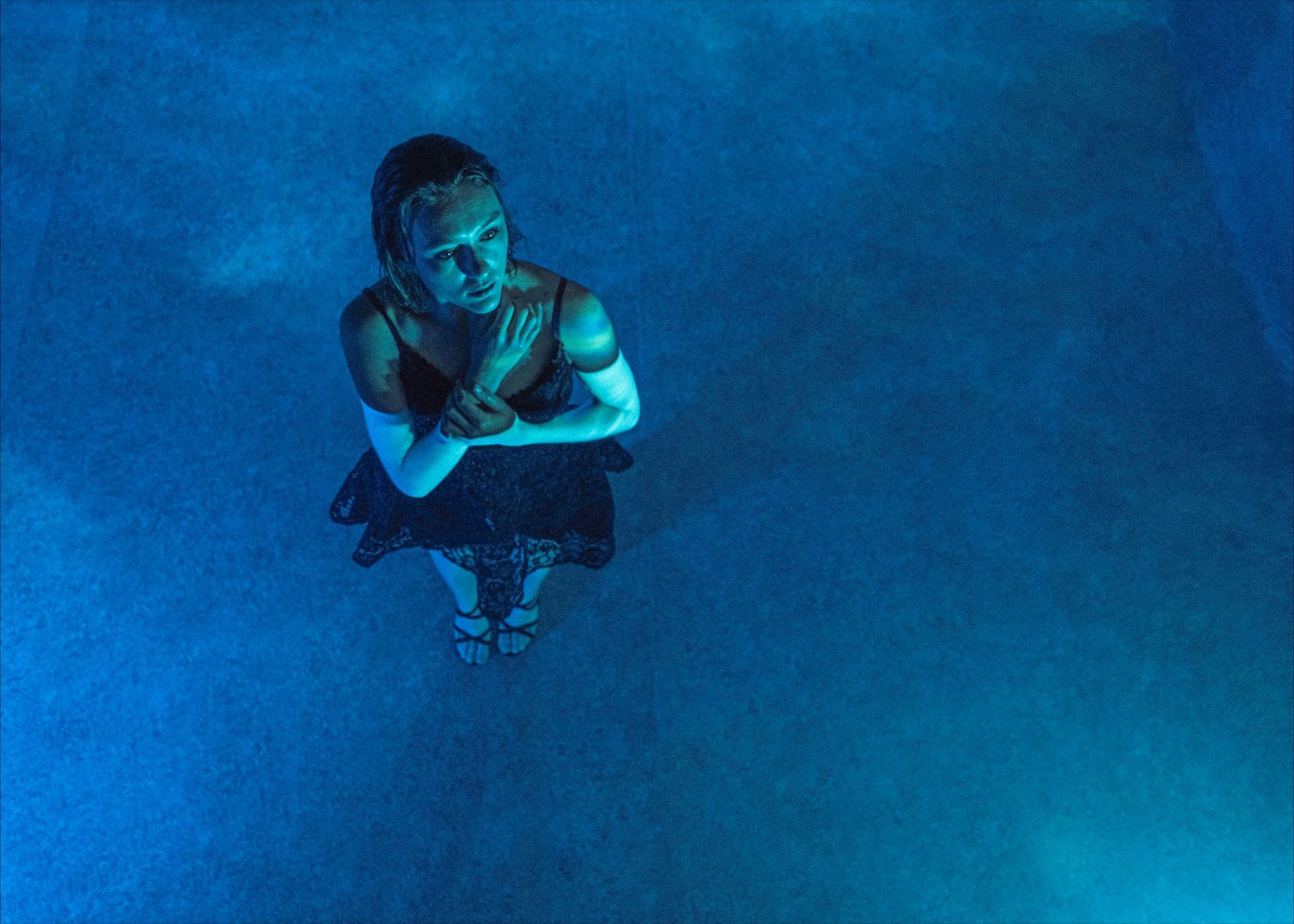 Woman in dark dress standing in blue-lit water with arms crossed, looking up.