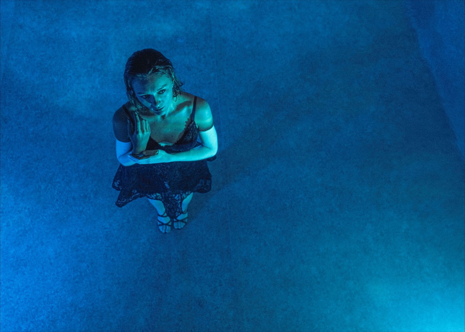 Woman in a black dress looking up with hands crossed, standing in blue-lit mist.