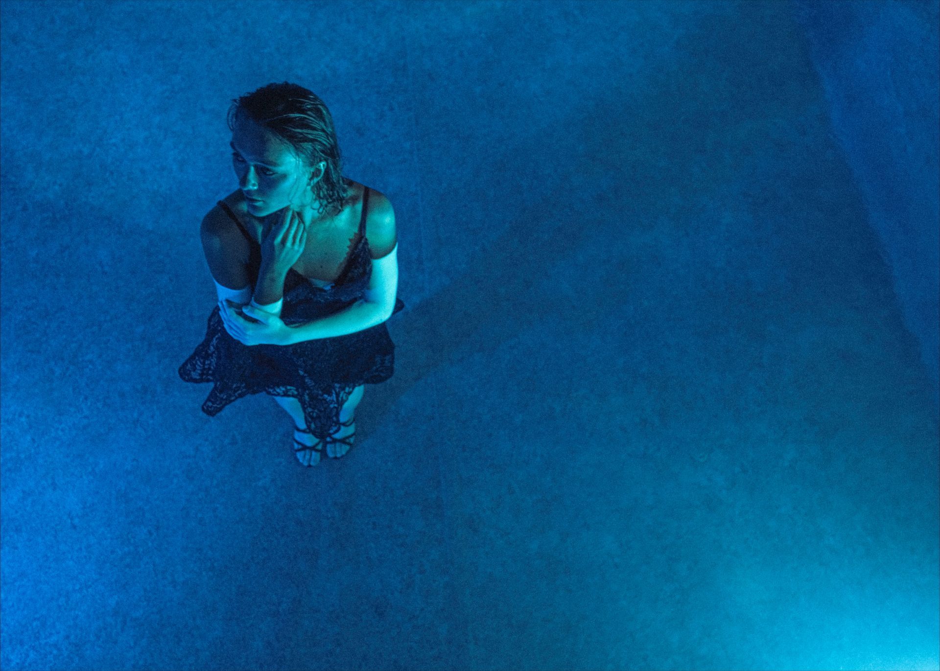 Woman in a dress, standing in blue-lit water, looking pensive, hand to chin.