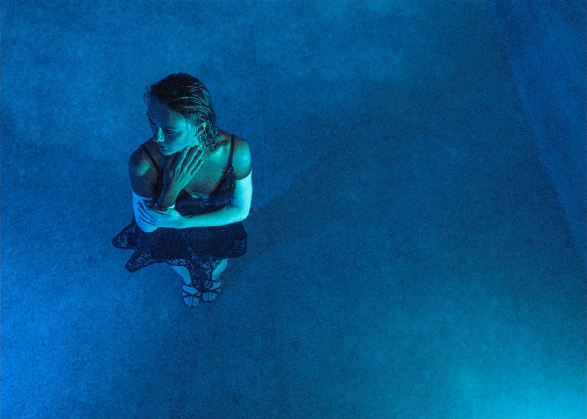 Woman in a black dress stands in blue light, arms crossed over chest.