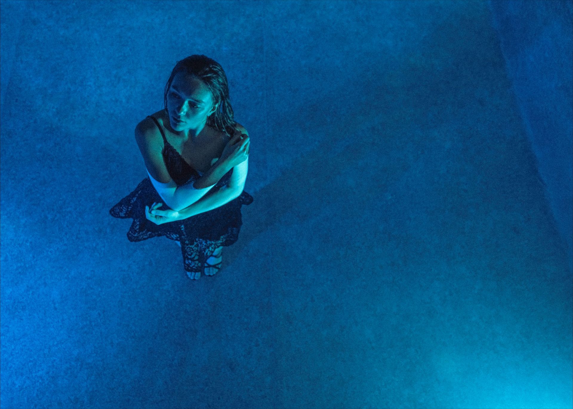 Woman in a dark dress, arms crossed, stands in blue-lit water, looking upward.