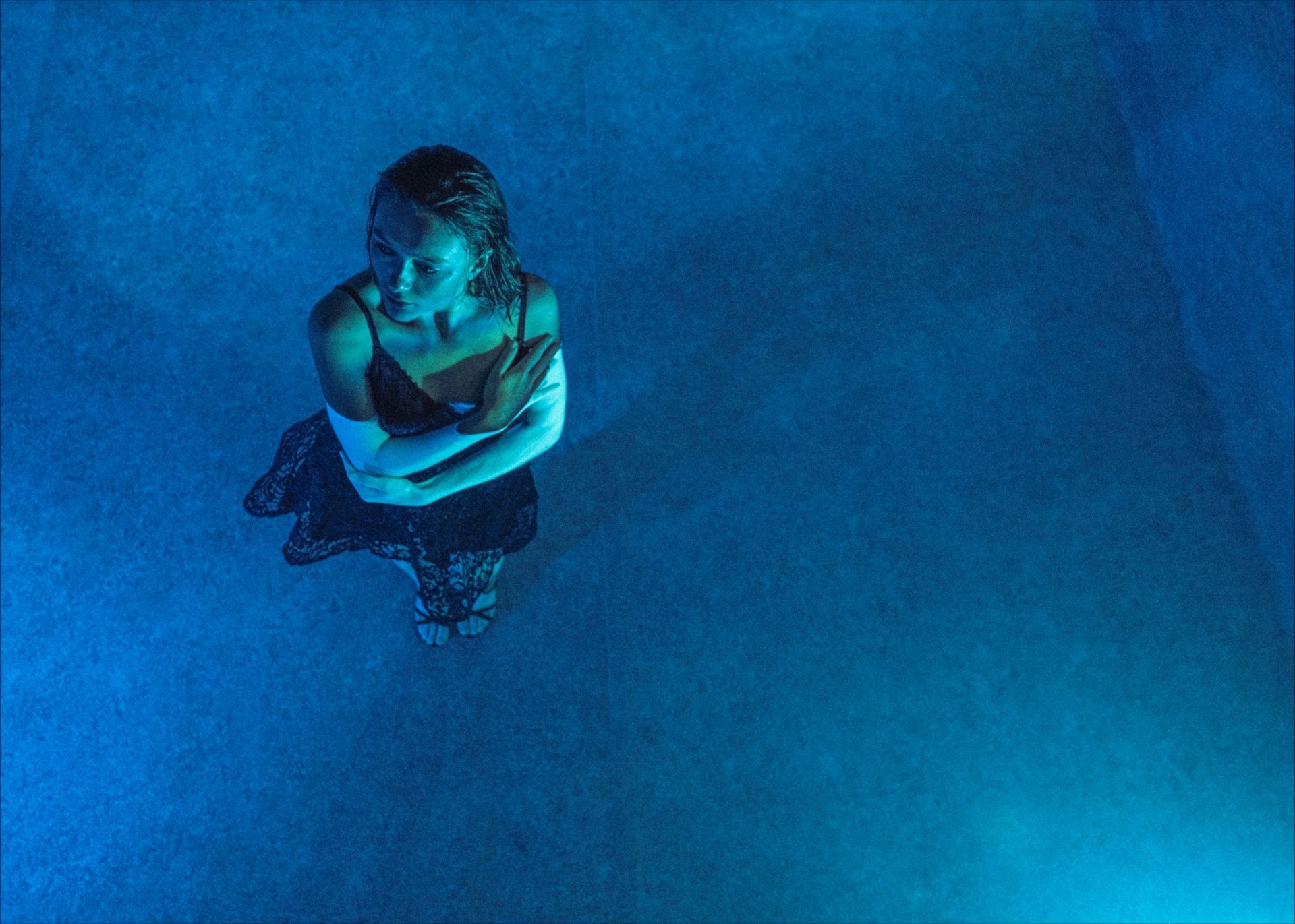 Woman in a dark dress, arms crossed, in a blue-lit pool.