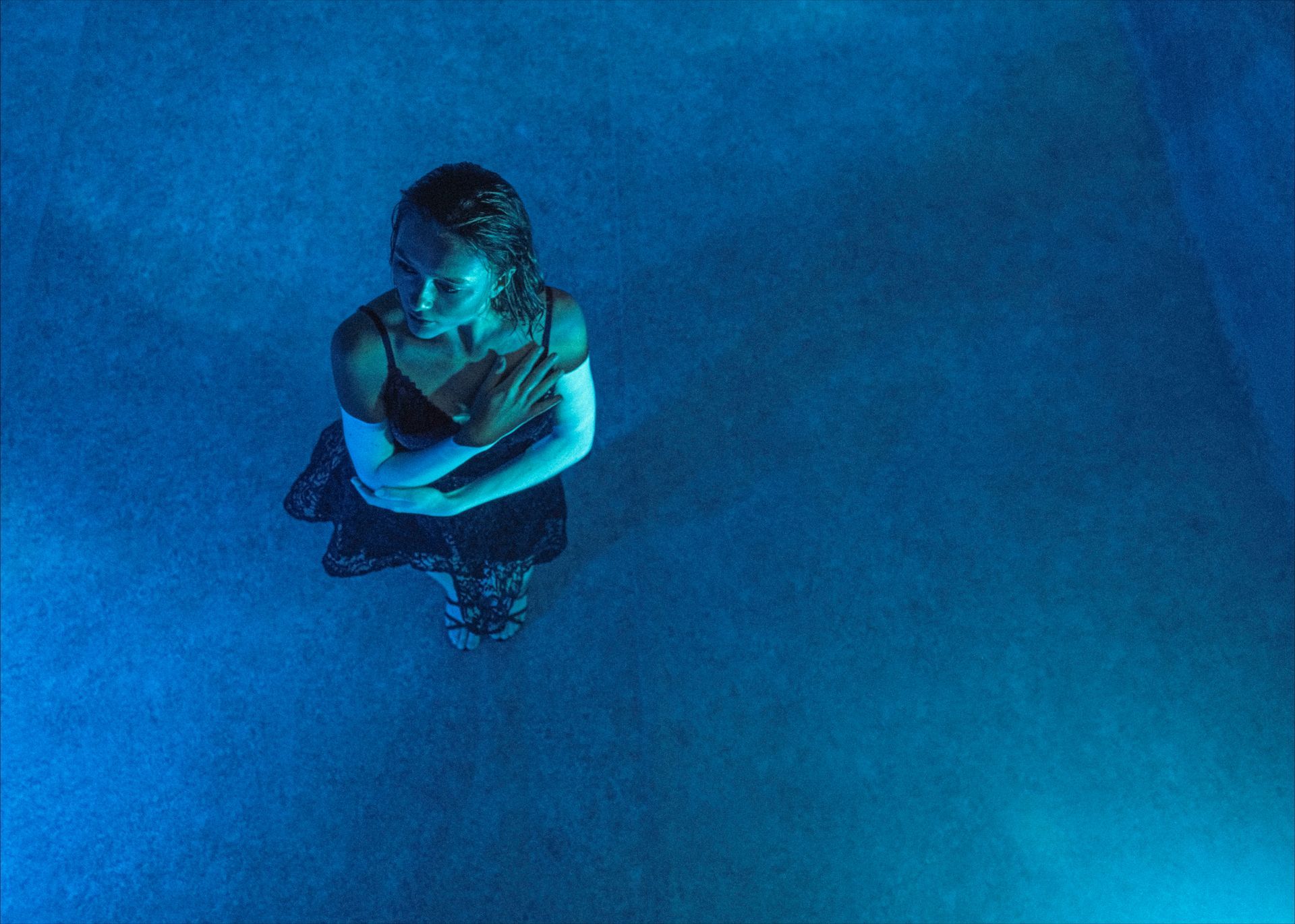 Woman in black dress, standing in blue-lit water with arms crossed over chest, looking upward.