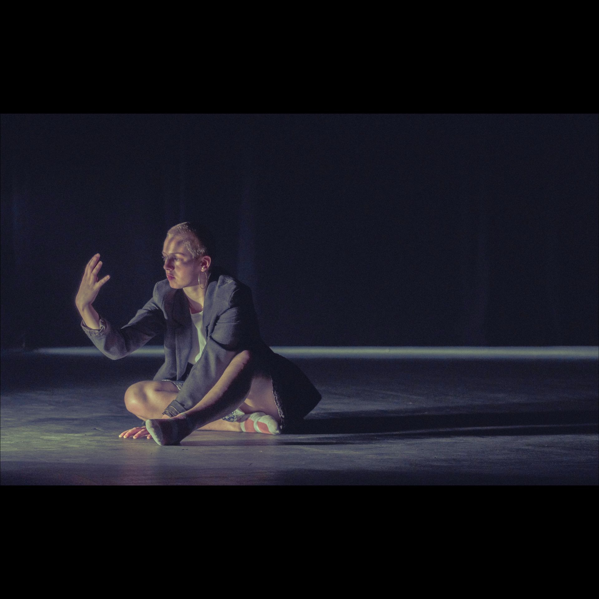 Person in suit jacket sitting on stage, arms raised, lit by spotlight.