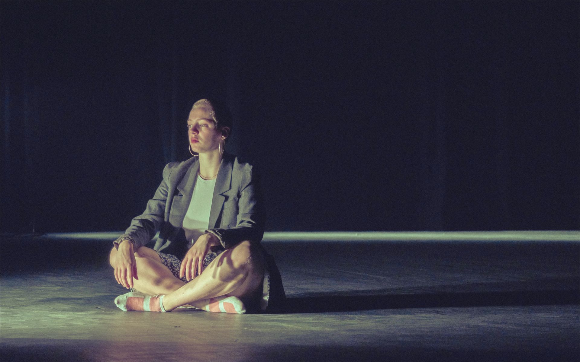Woman in a blazer sits cross-legged, eyes closed, in a dimly lit space. Spotlight shines on her.