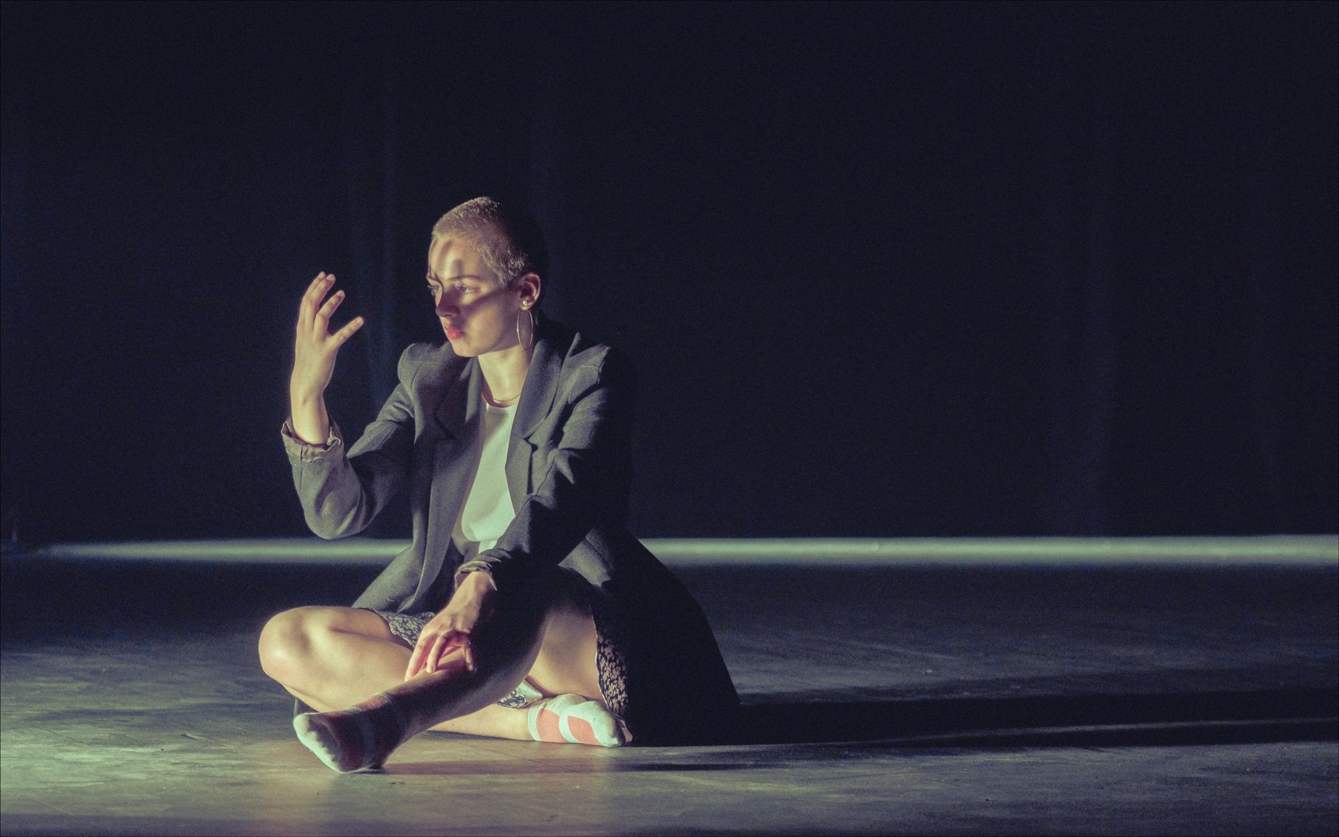 Woman in blazer sits cross-legged, hand raised, in spotlight on dark stage.