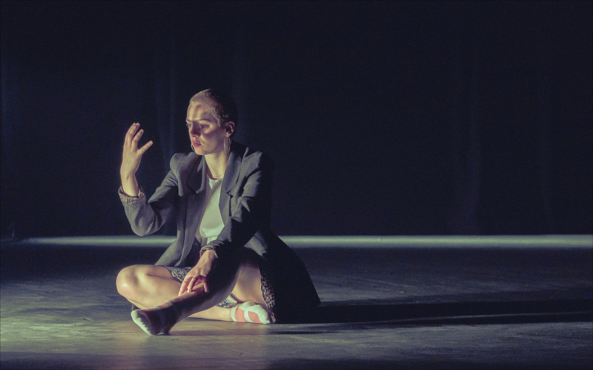 Person in blazer sits cross-legged, hand raised, in spotlight. Dark background.