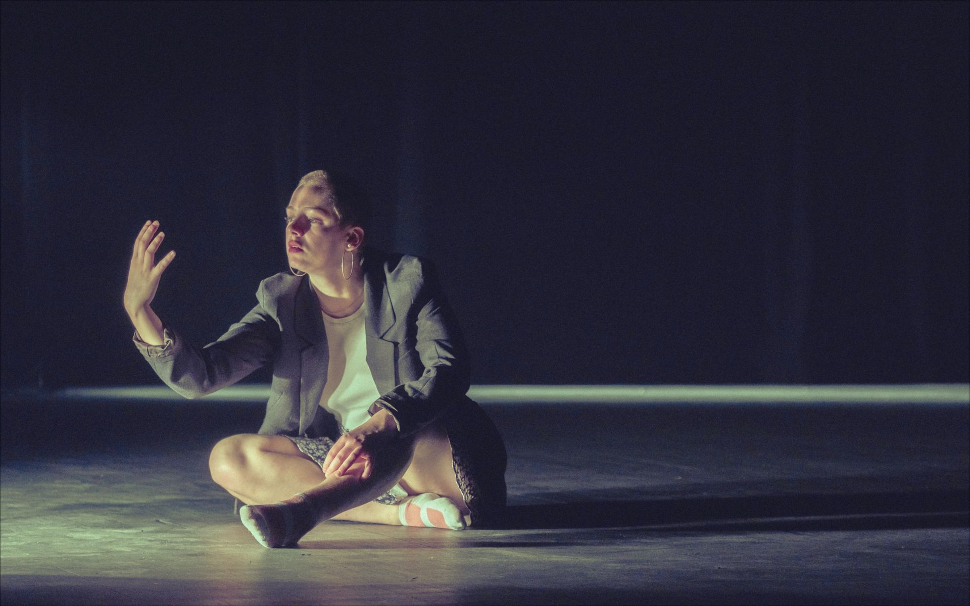 Person seated, cross-legged, on a dark stage; arm outstretched, lit from above; wearing a blazer.