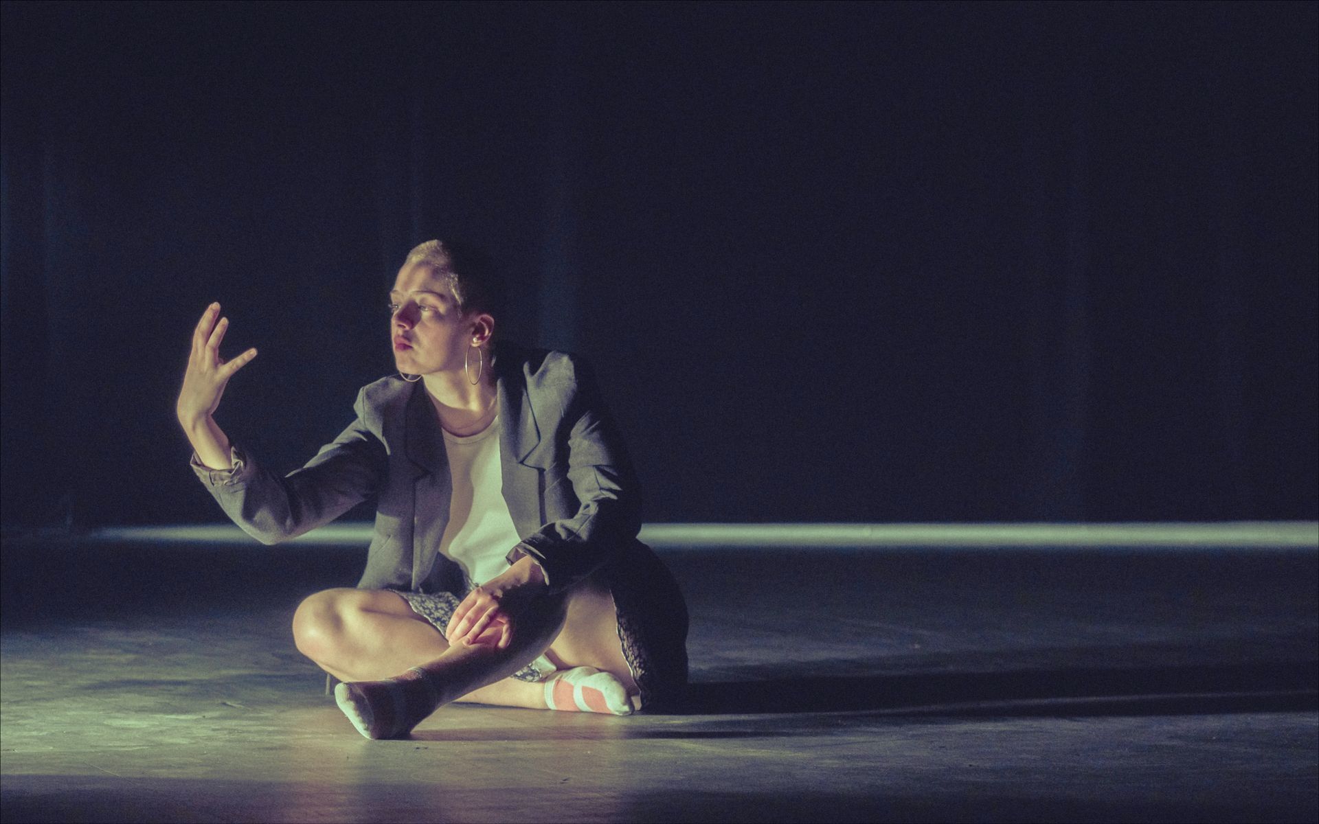 Person sitting cross-legged, arm raised, on a stage lit with shadows.