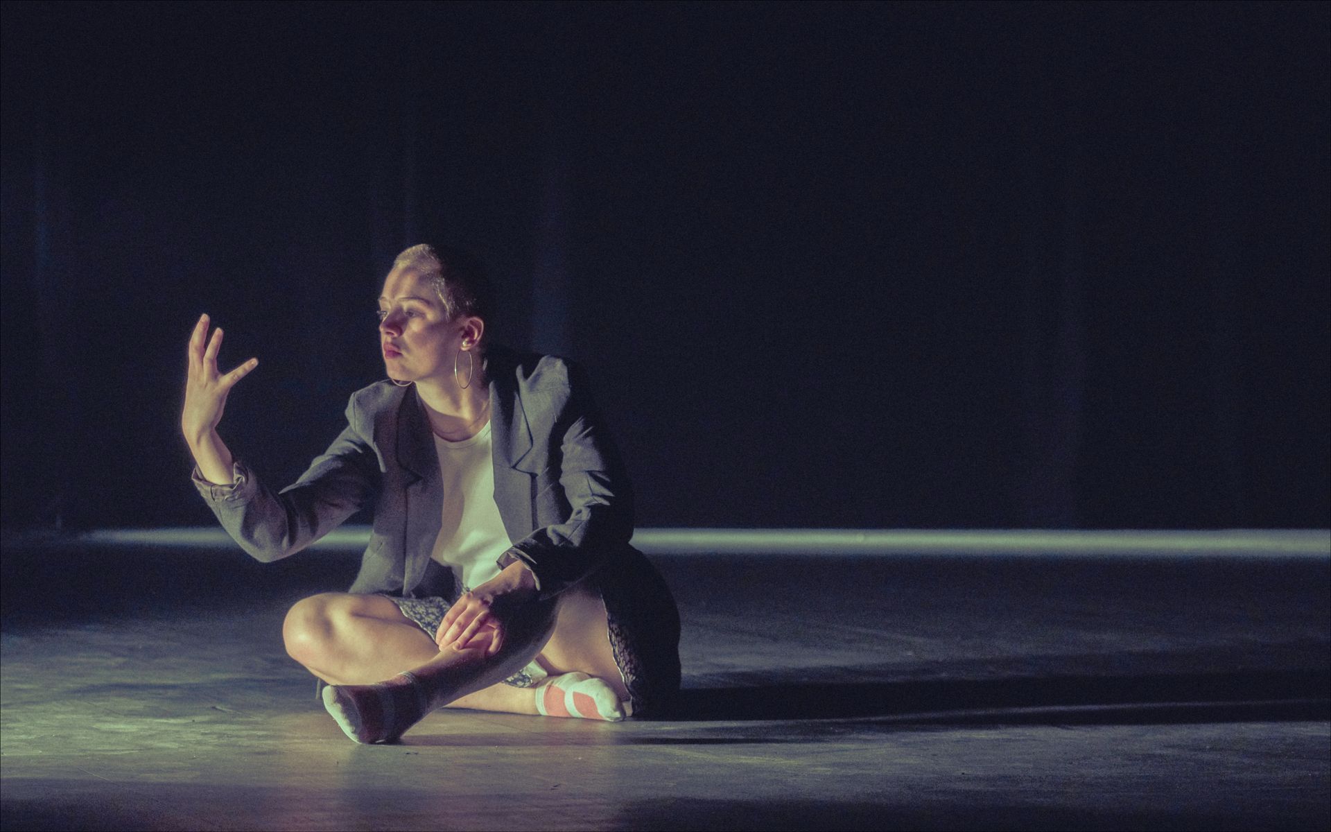 Person sitting cross-legged, illuminated in dark space, gesturing with hand raised.