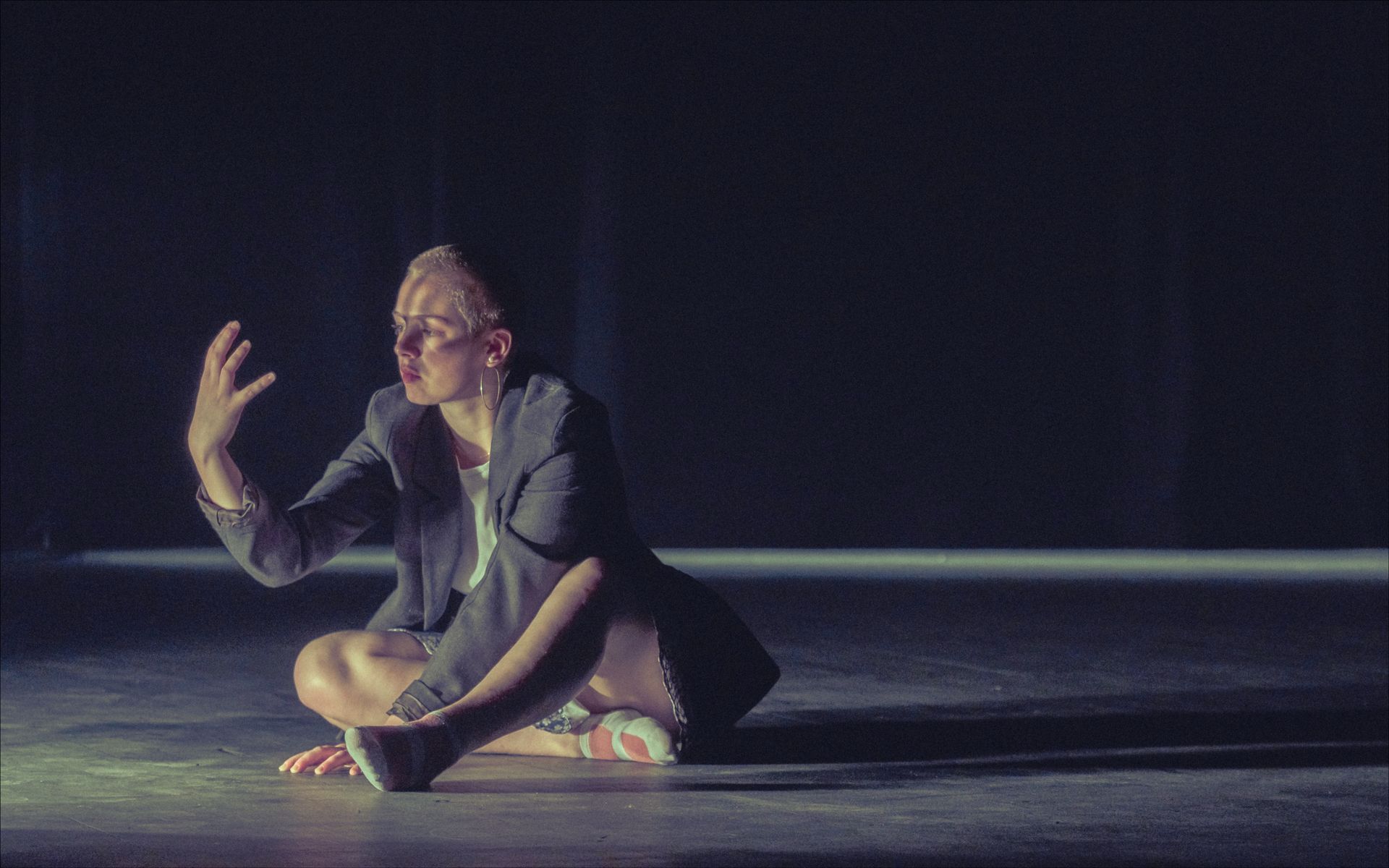 Person seated on floor, arms outstretched, wearing jacket, lit by stage lighting.