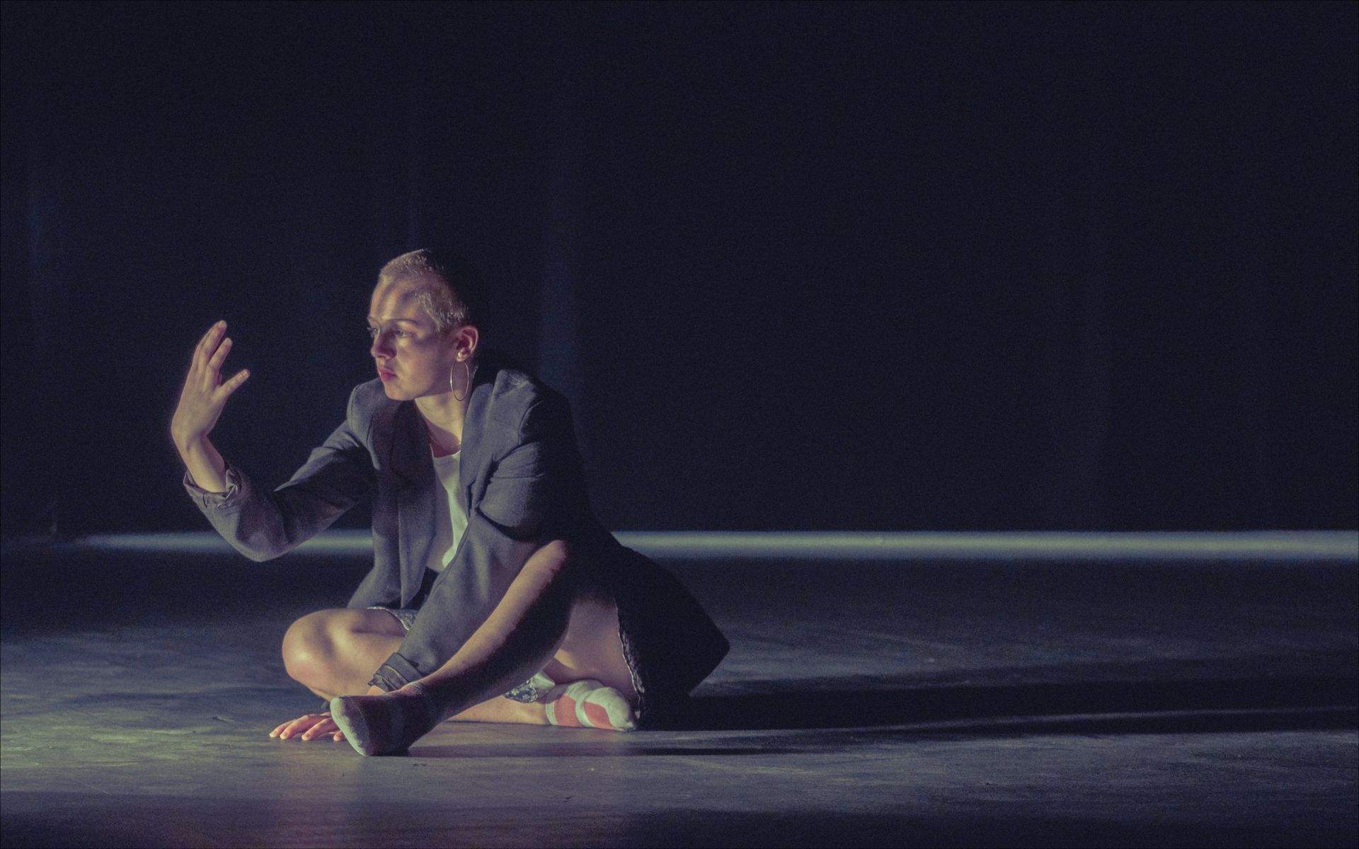 Person in a blazer sits on the floor with legs crossed, arm raised. Dark setting, lit by a single light.