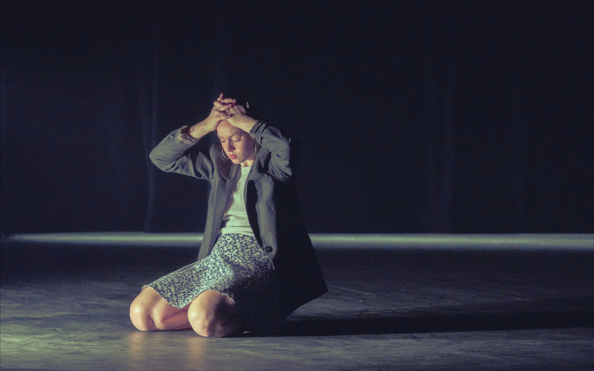 Person kneeling on stage, hands on head, looking distressed. Dark background, spotlight.