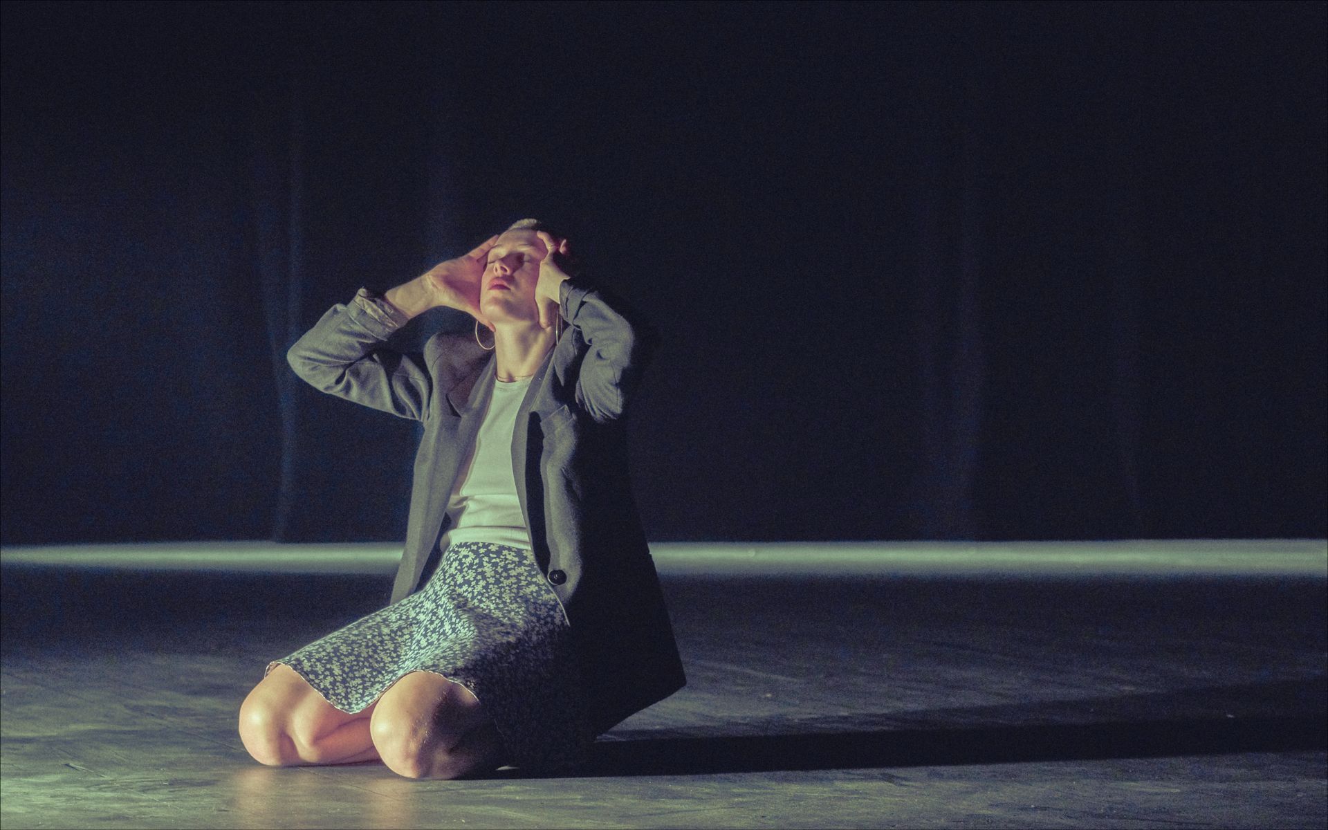 Person kneels, hands to head, lit stage. Dark background, wearing coat and patterned skirt.
