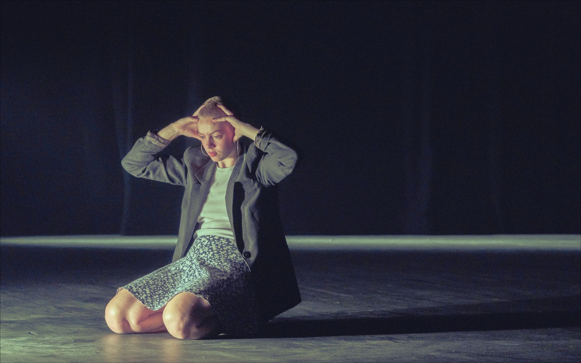 Person kneels, hands to head, dark setting.  Gray coat, patterned shorts.  Stage spotlight.