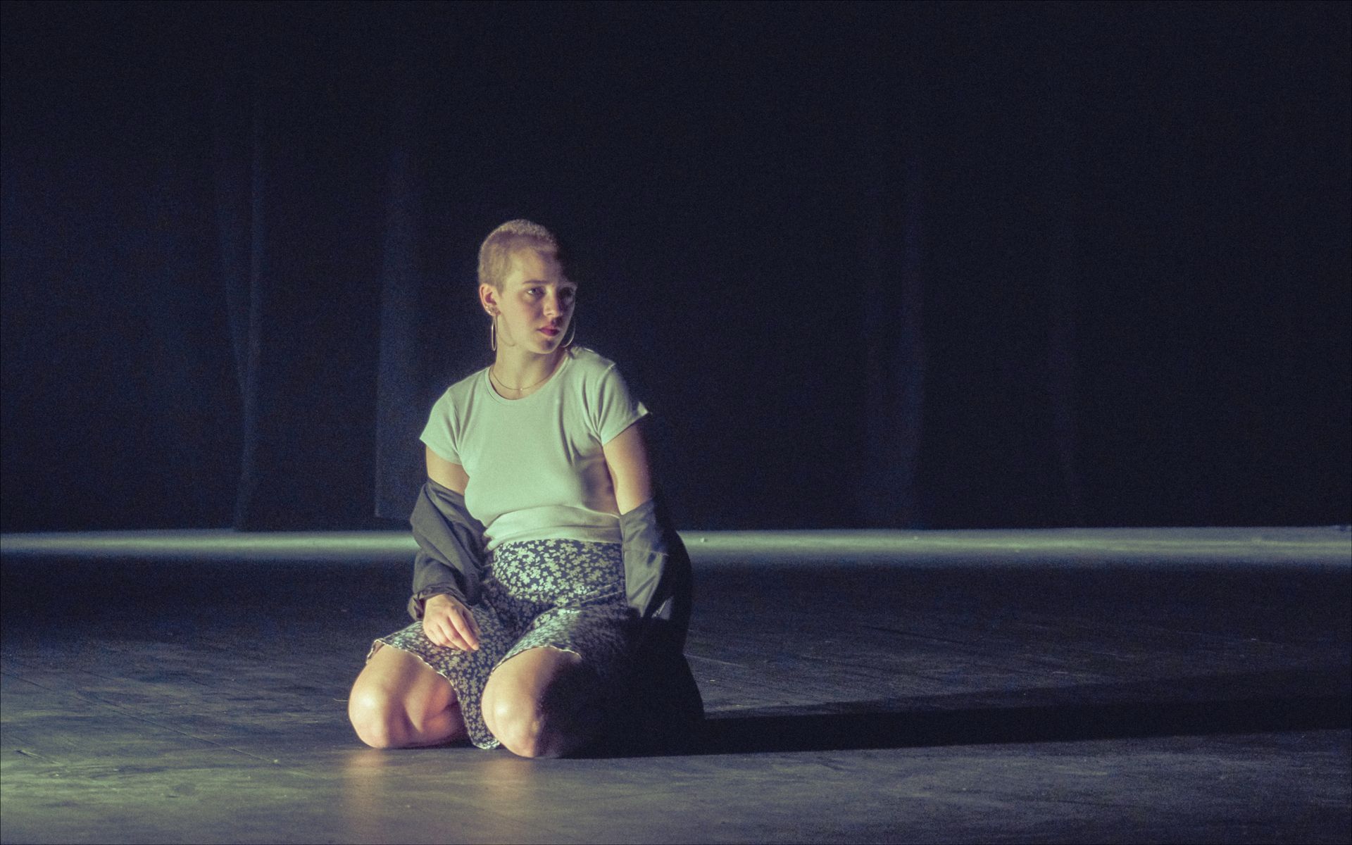 Person kneels on stage, looking left, jacket on shoulders. Dark stage.