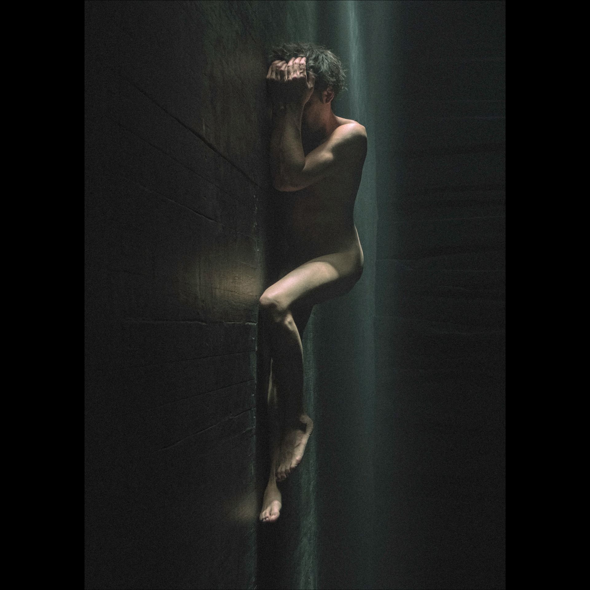 Naked person clinging to wall in dimly lit space, legs crossed, arms over head. Light shines from above.