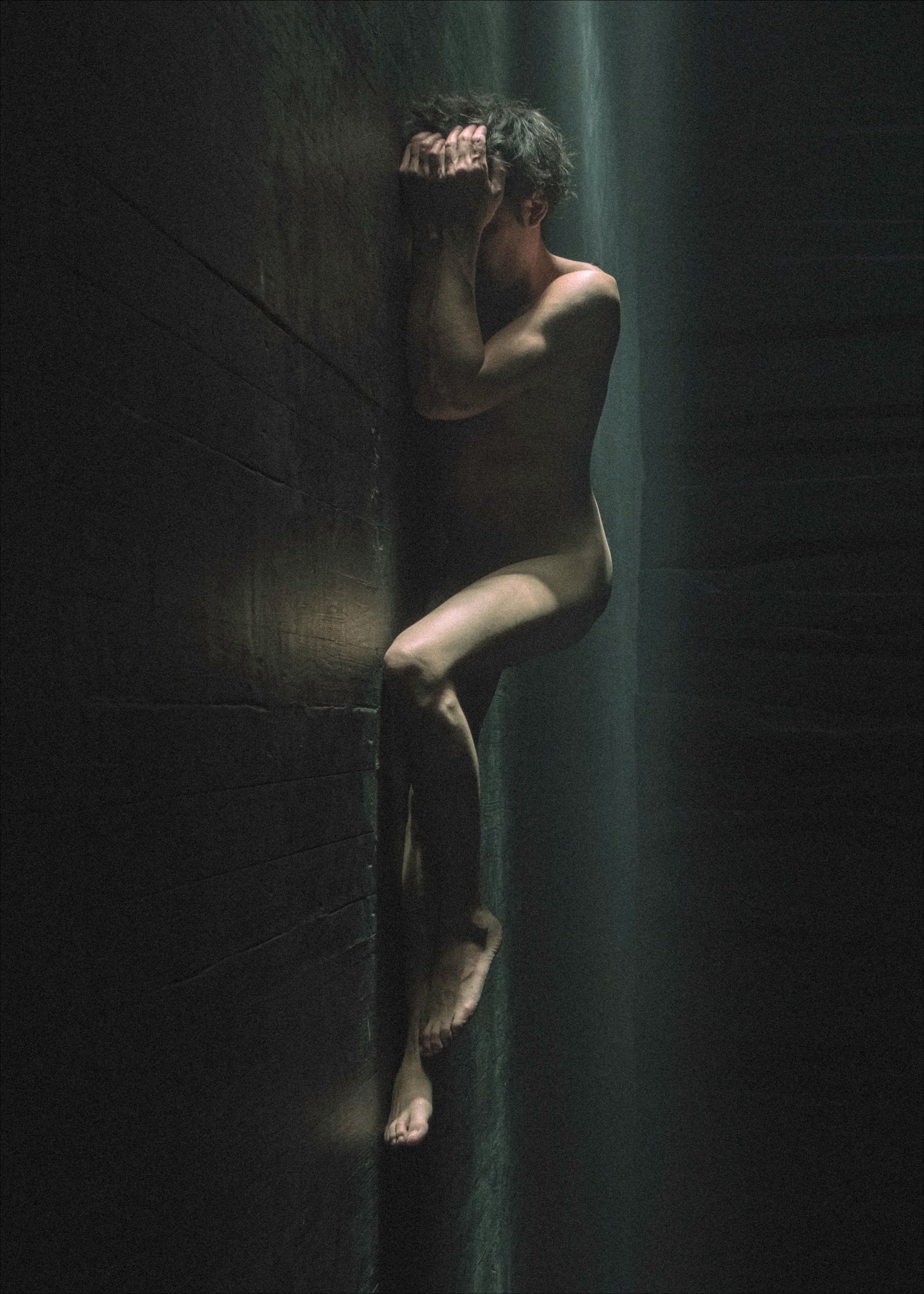 Nude figure pressed against a dark wall, arms covering face, legs bent, a shaft of light illuminates the body.