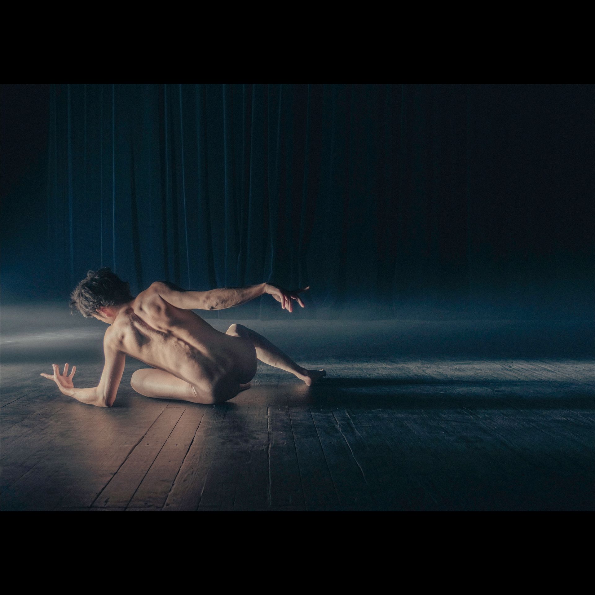 Naked dancer on wooden floor, backlit by dark curtains, in a pose of movement.