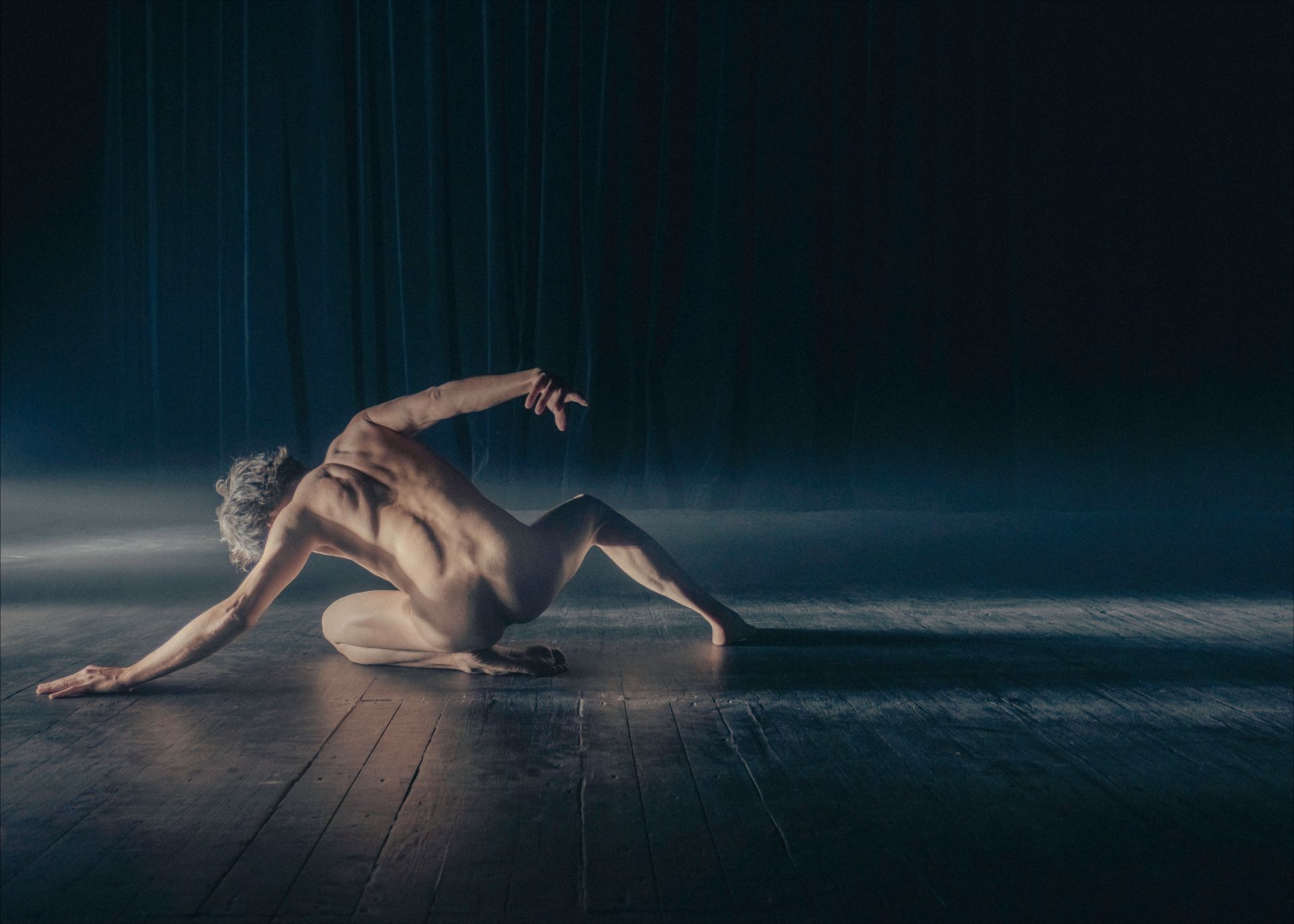 Nude dancer on stage, bent over with arm outstretched, back to viewer. Dark, moody lighting.