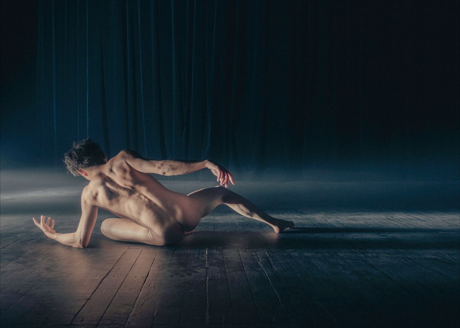 Naked person, back to viewer, on wooden floor, reaching out, performing dance. Dark stage with blue curtain.
