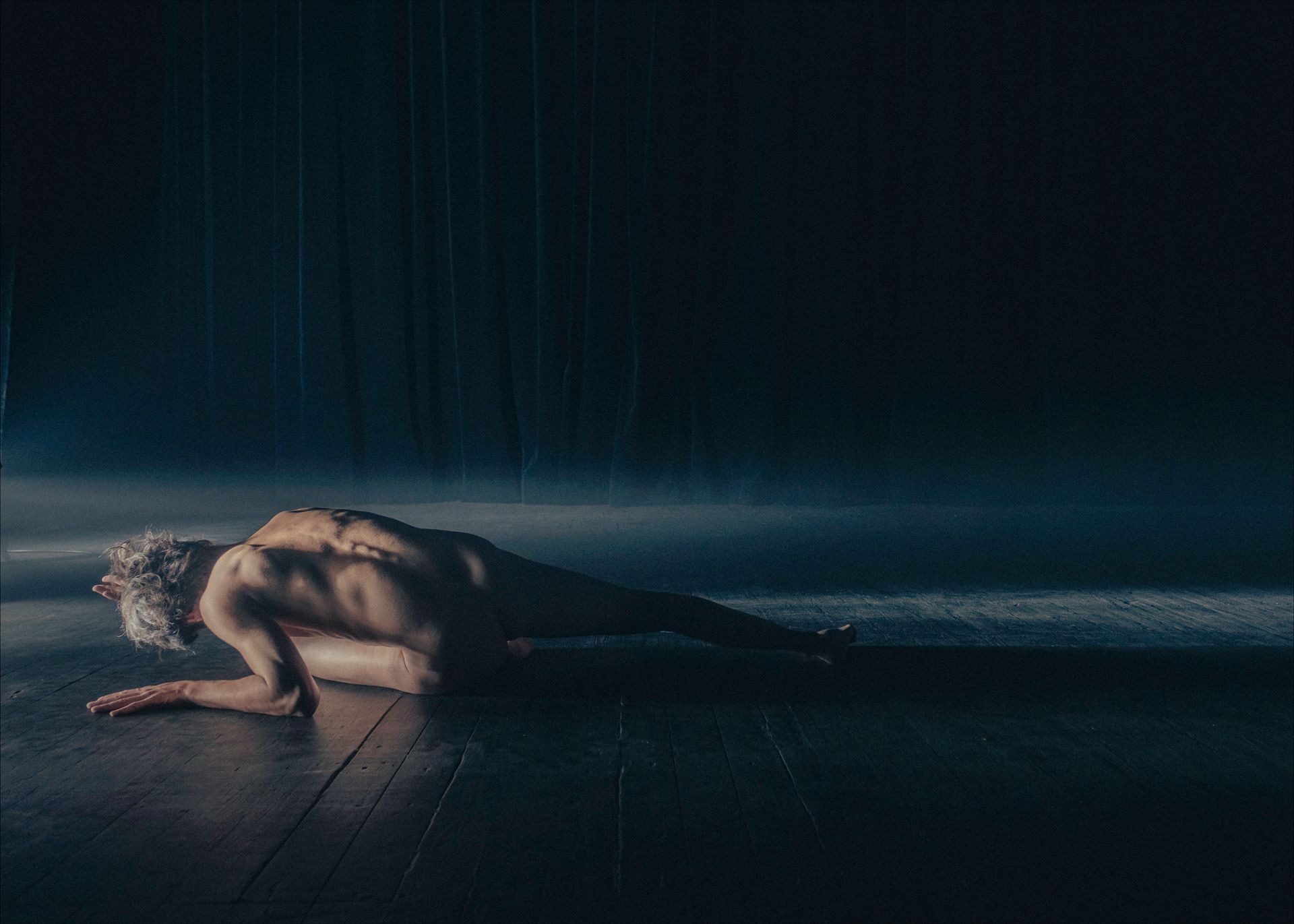 A person crouched on the floor in a dim room; dimly lit, side profile.