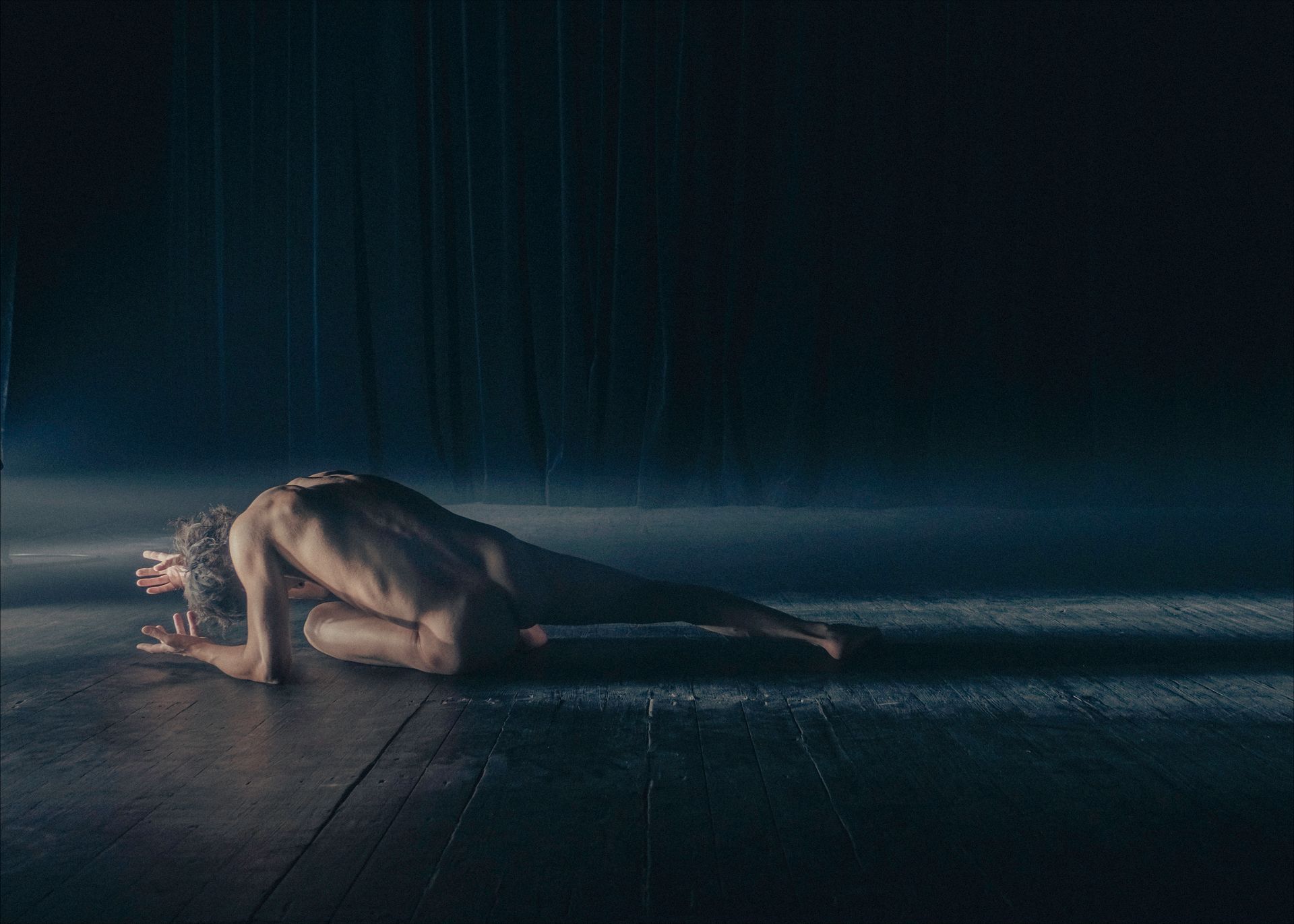 A person on a dark stage, bent over, seemingly in pain, with droplets falling.