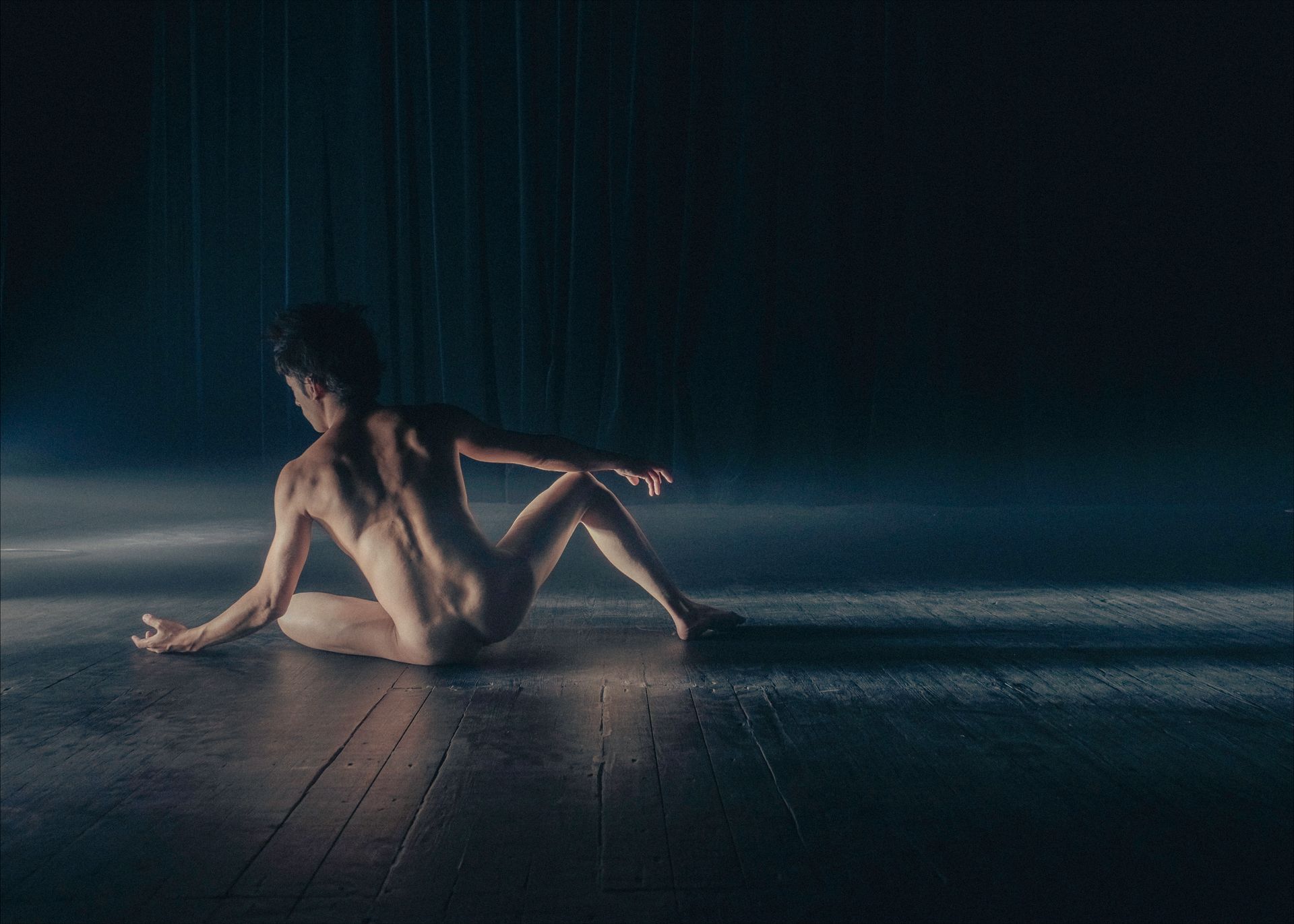 Nude person sits on a dark, wet floor, arms outstretched, back to viewer. Blue and white lighting.