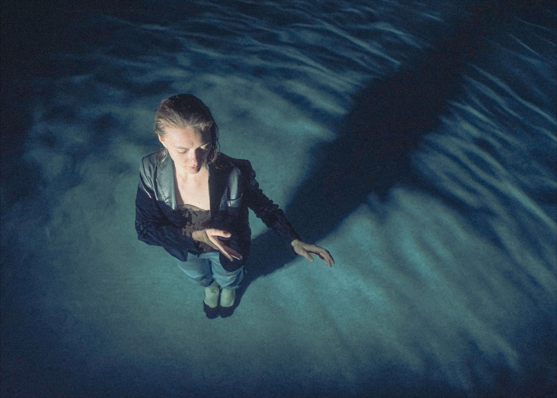 Woman in dark clothes stands in water, reaching out, shadow on blue surface.