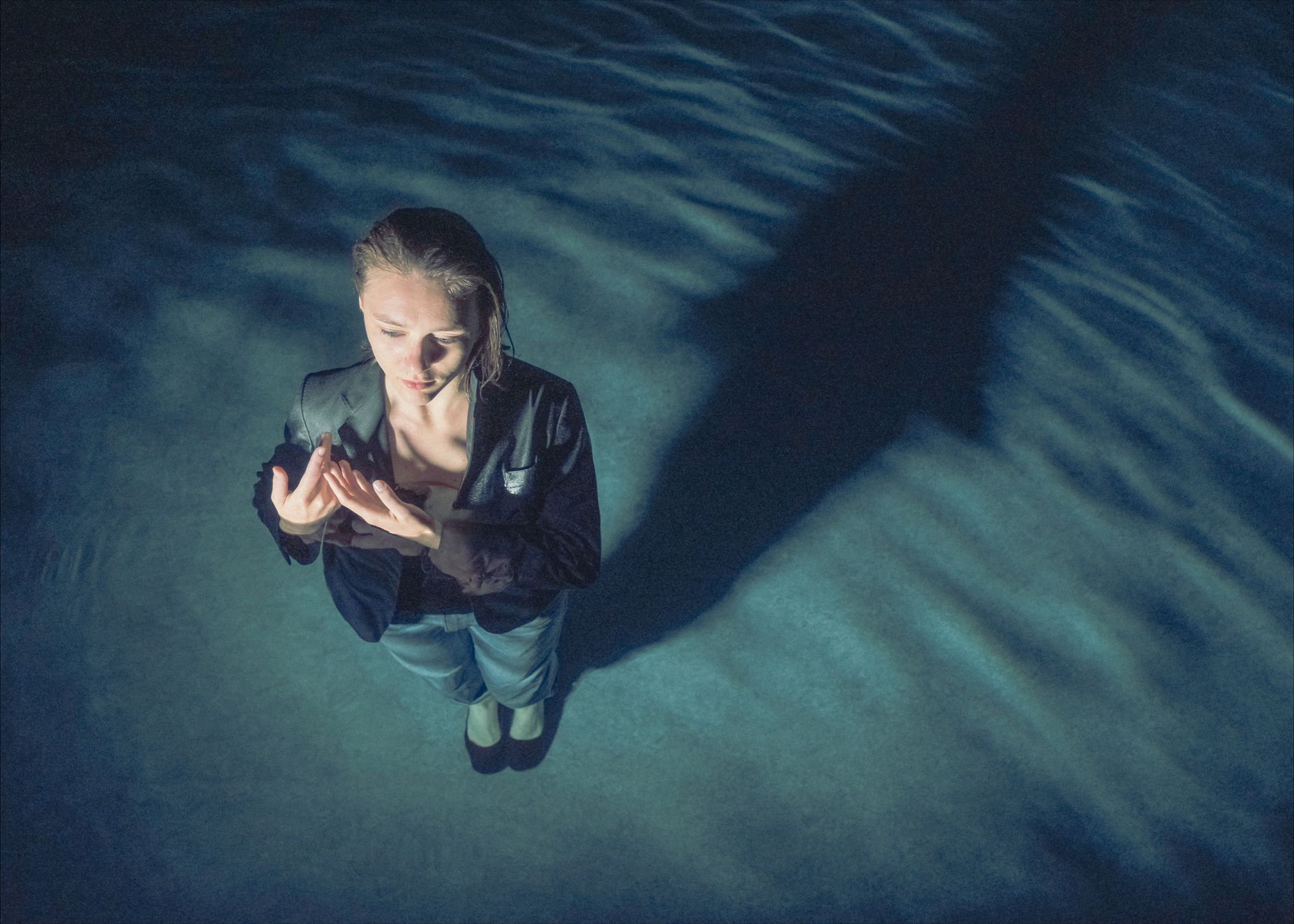 Woman in jacket and jeans, arms raised, casting shadow on blue rippled surface.