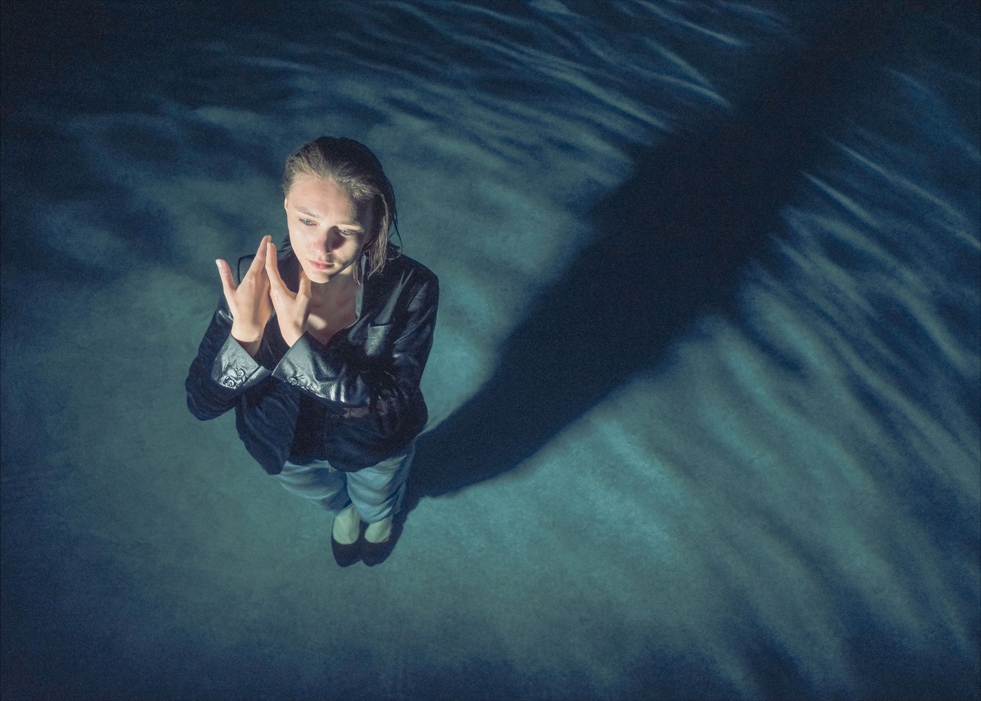 Woman, hands clasped, stands in blue-toned water, long shadow stretches beside.
