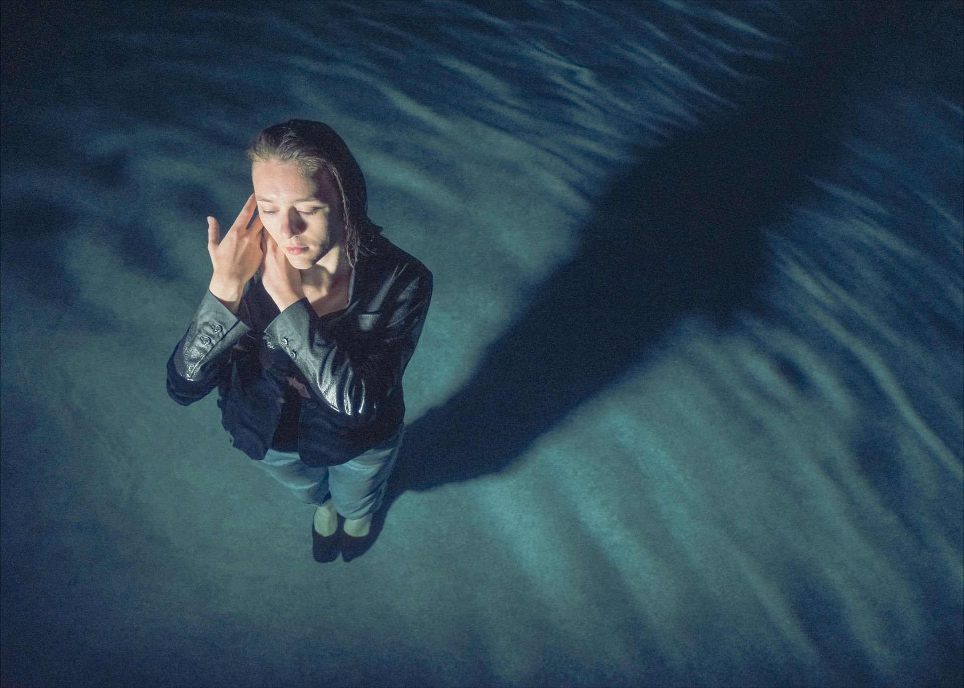 Woman in wet clothes stands in water, hands to her ears, long shadow.