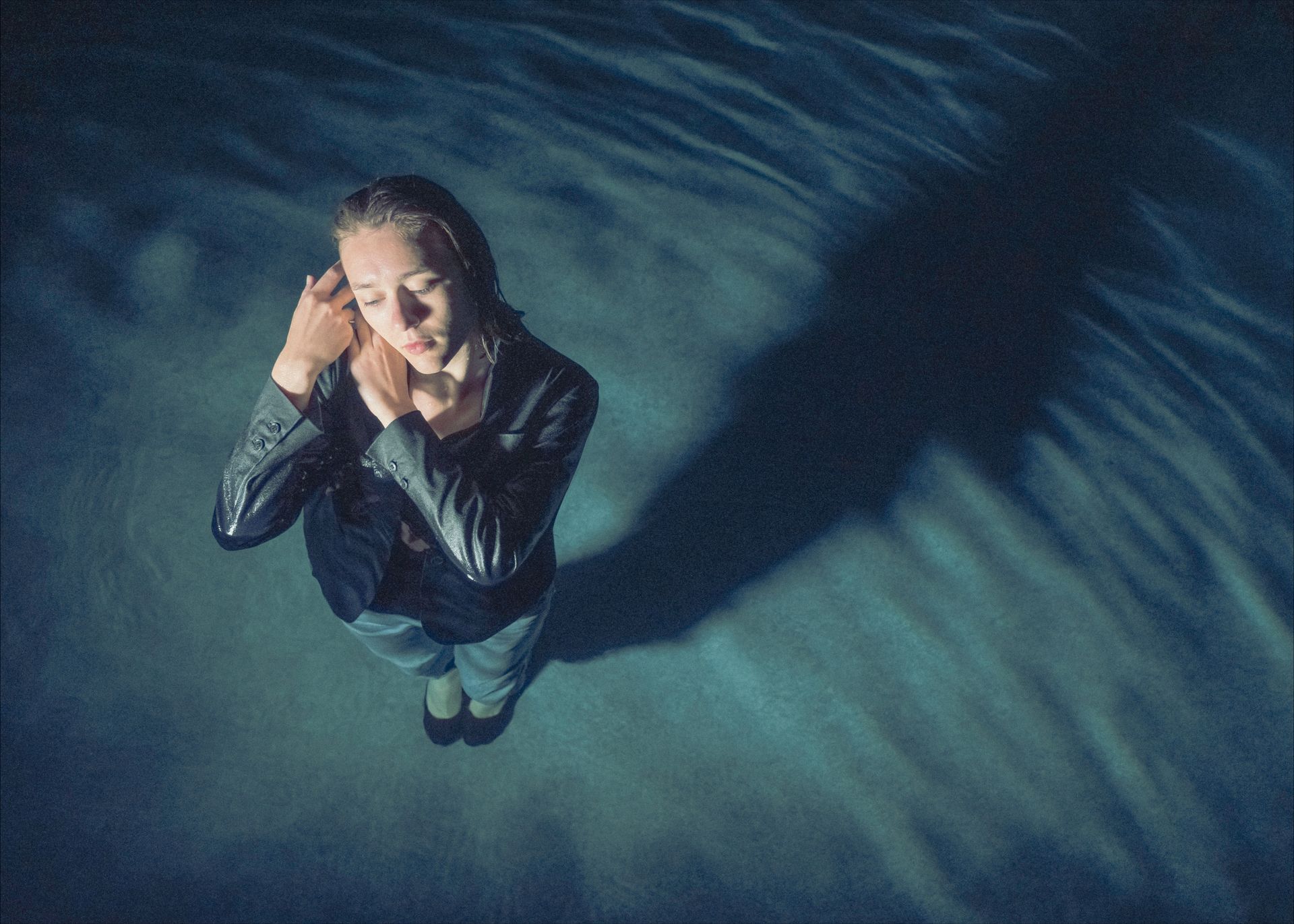 Woman standing in water, arms raised to head; long shadow; blue tones.