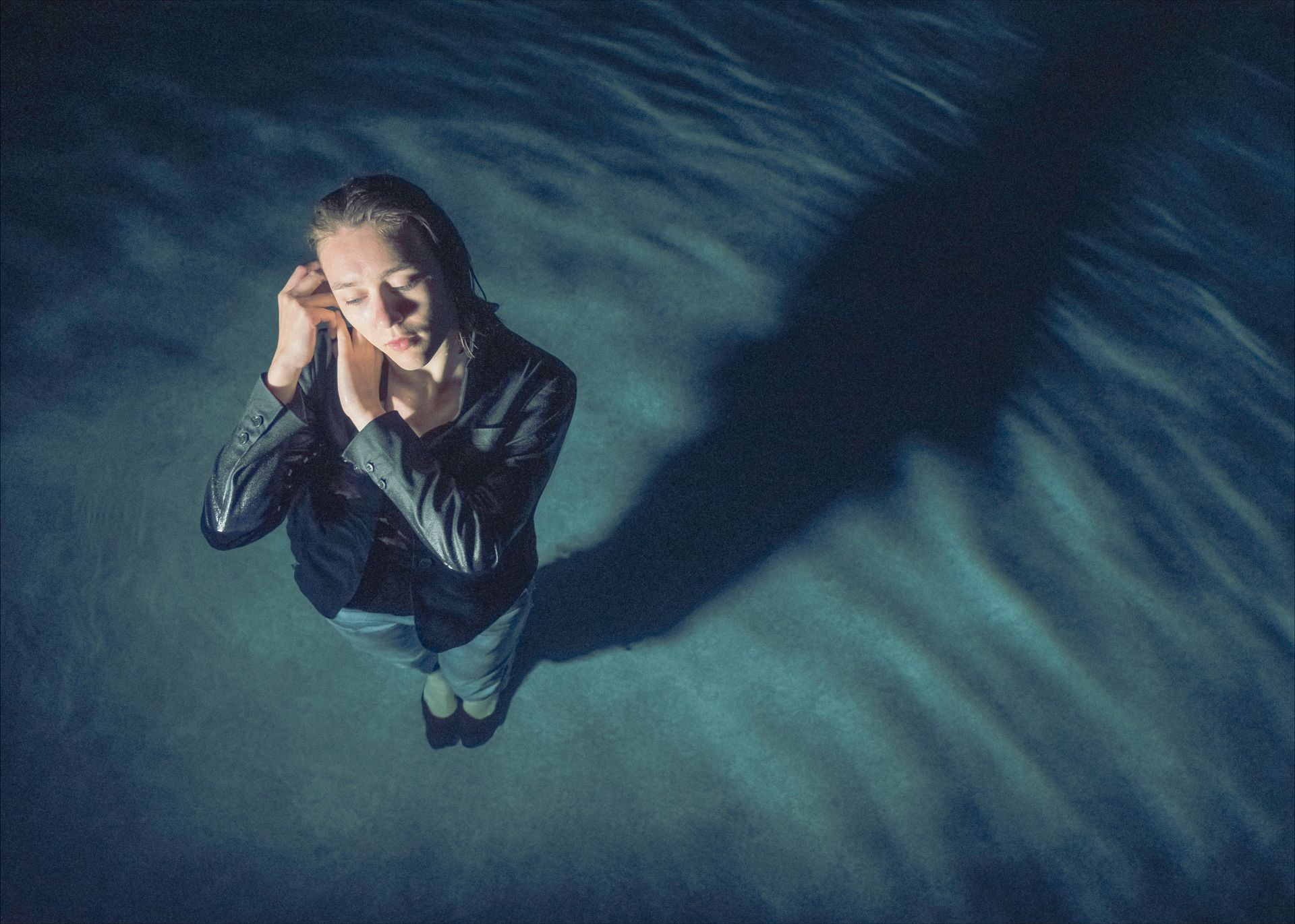 Woman in black jacket, eyes closed, hands near ears, standing in blue-lit space with long shadow.