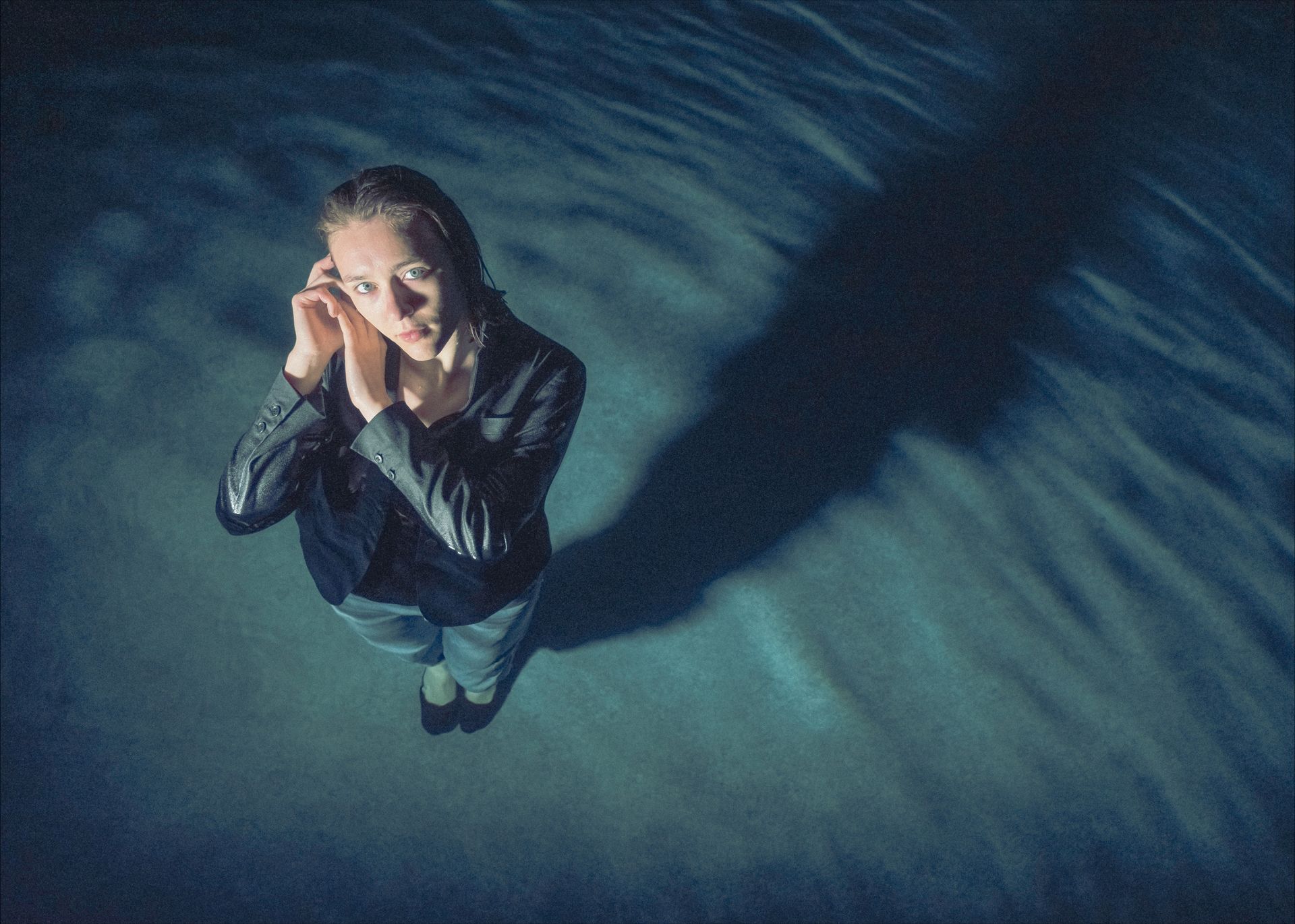 Woman in water, looking up, hands near ears, wearing dark clothing. Shadow on the water.