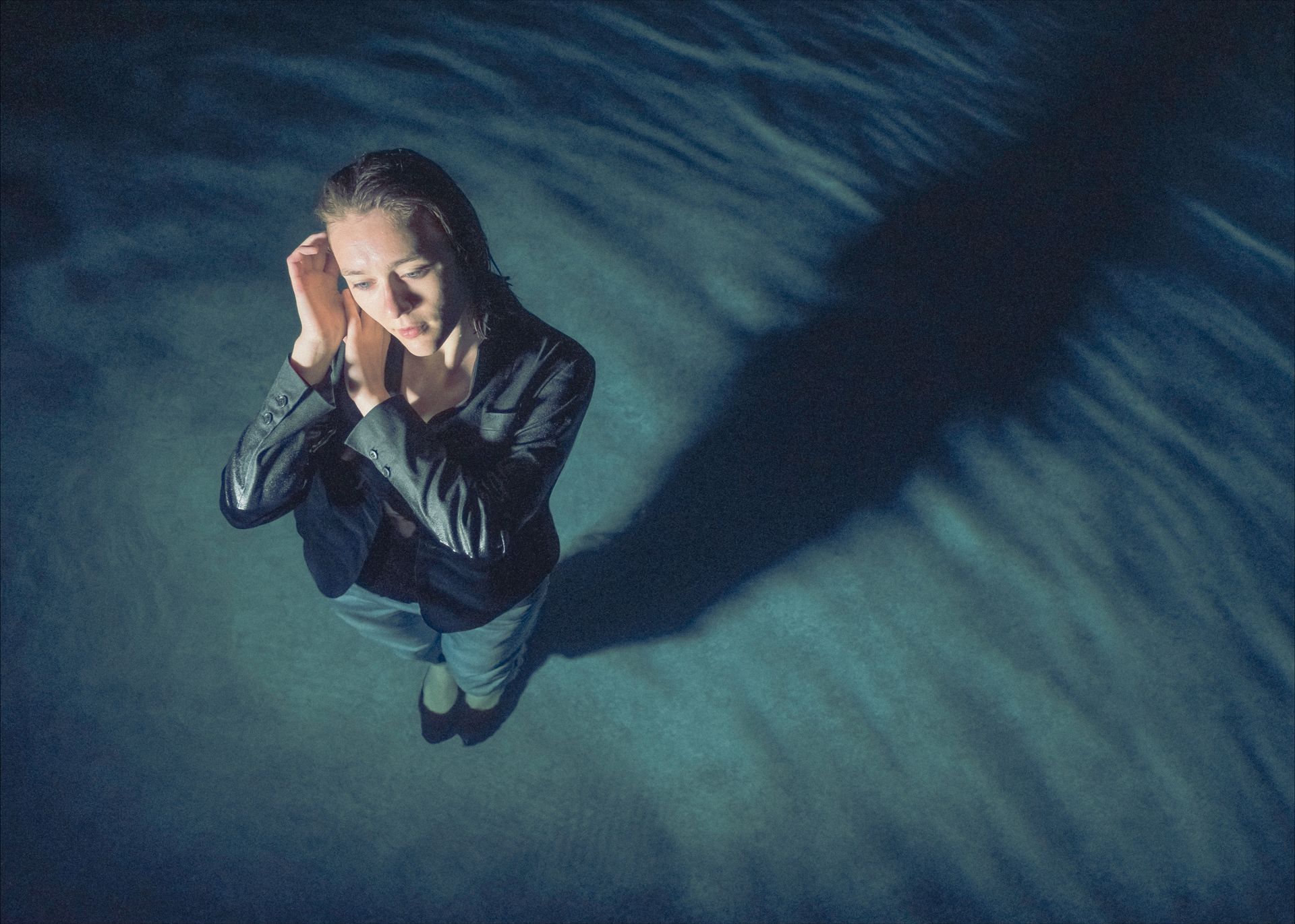 Woman in dark jacket and jeans, head bowed, hands near ears, standing in blue, wave-like setting.