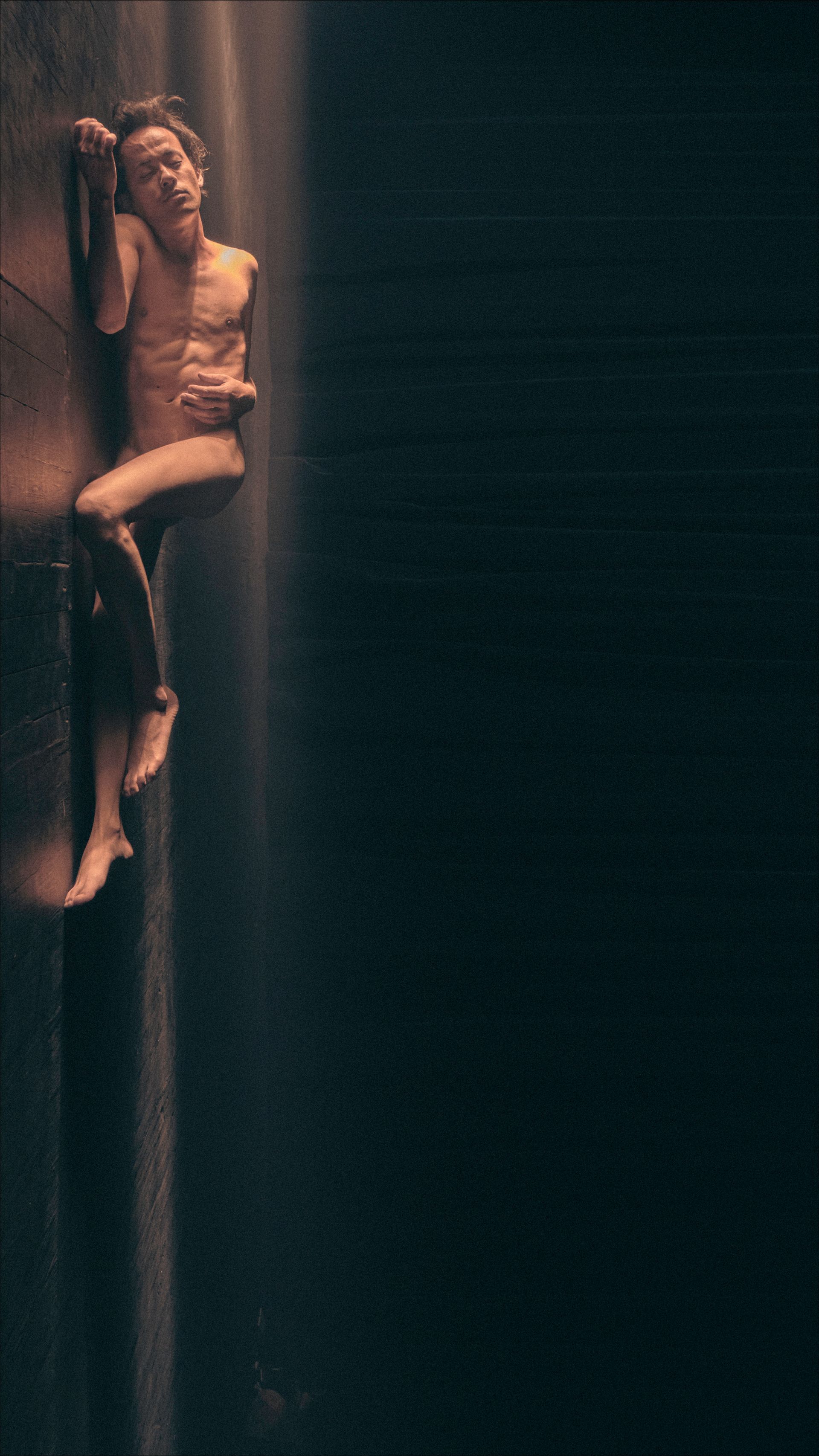 Man clinging to a dark wall, lit from above, bare chest, dark pants.