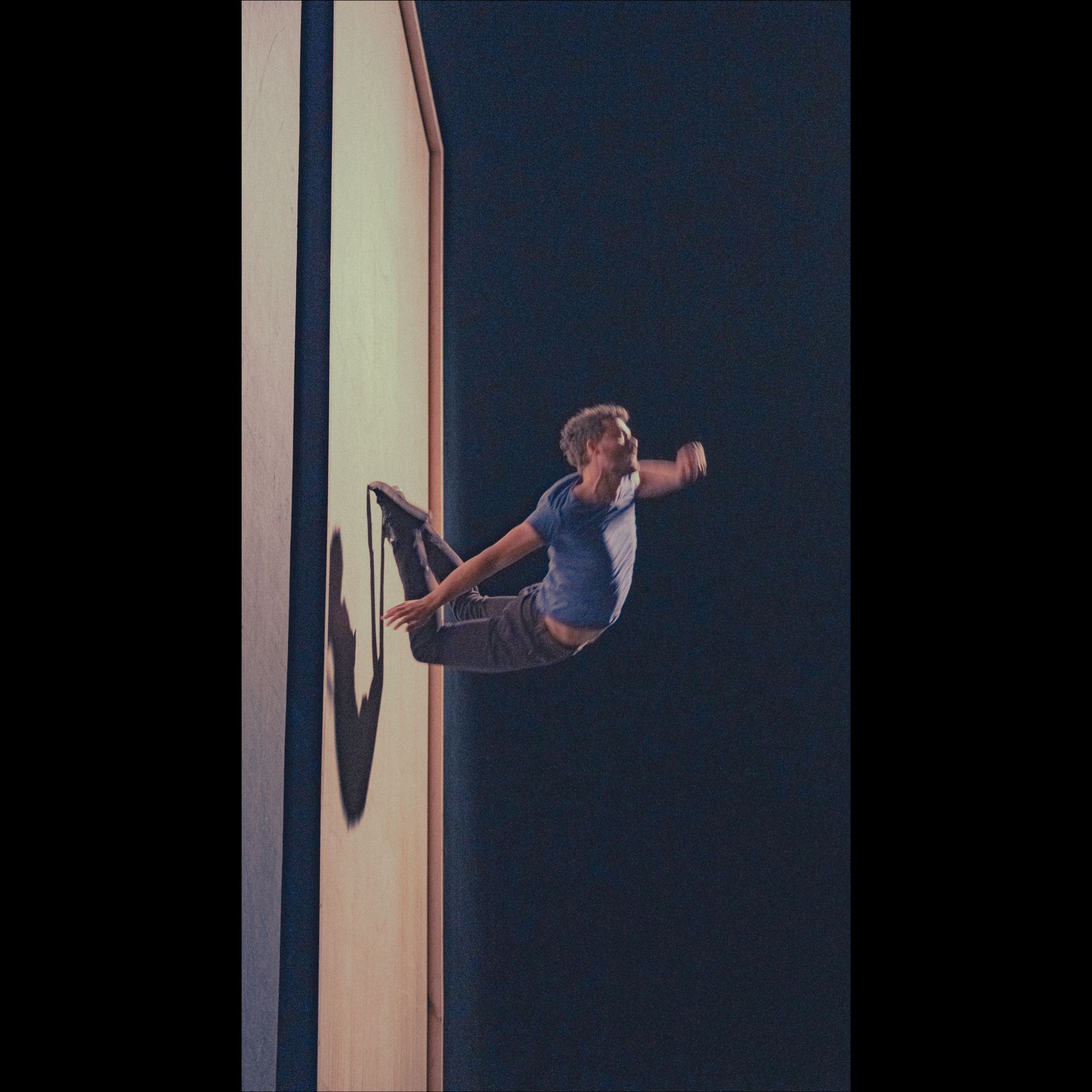 A man performs a mid-air acrobatic move, appearing to be pushing off a wall; dark background.