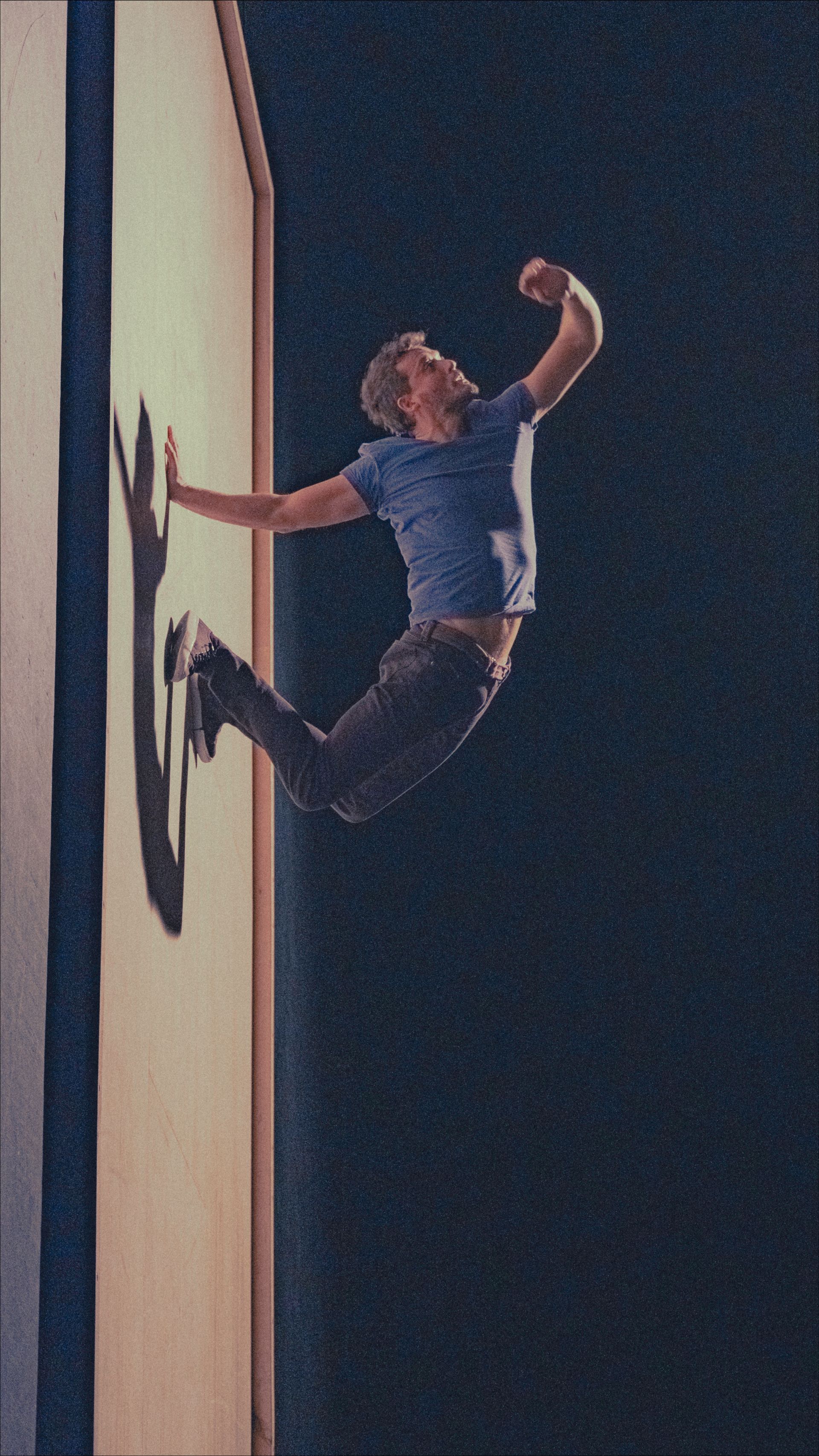 Man leaping off a wall, arm raised in triumph, with a long shadow against a dark background.