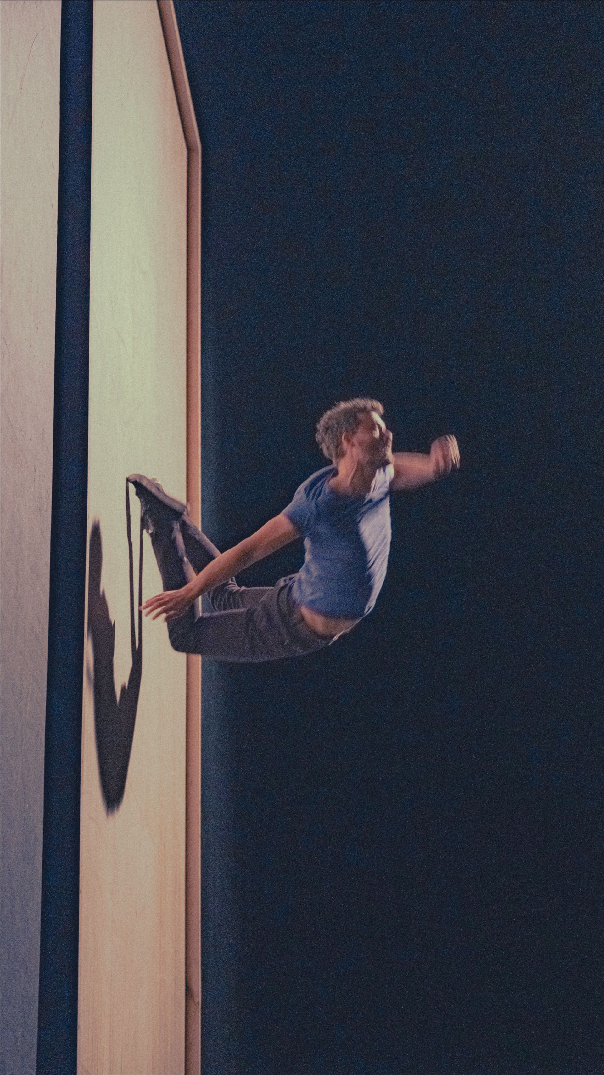 Man arches backward mid-air, grasping a wall. He is wearing a blue shirt and grey pants. Dark setting.