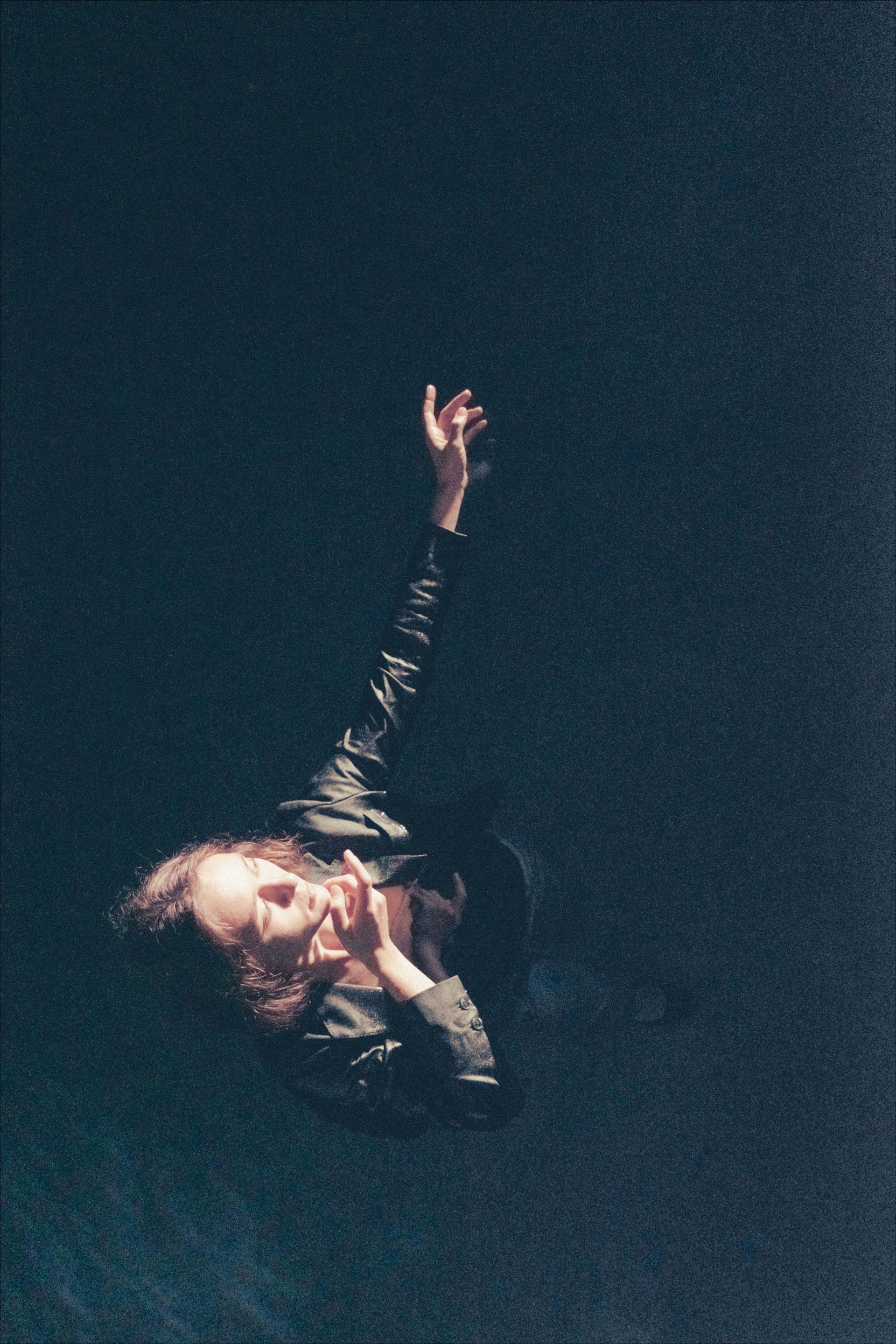 Person with long hair, dark jacket, reaching up, dark background.