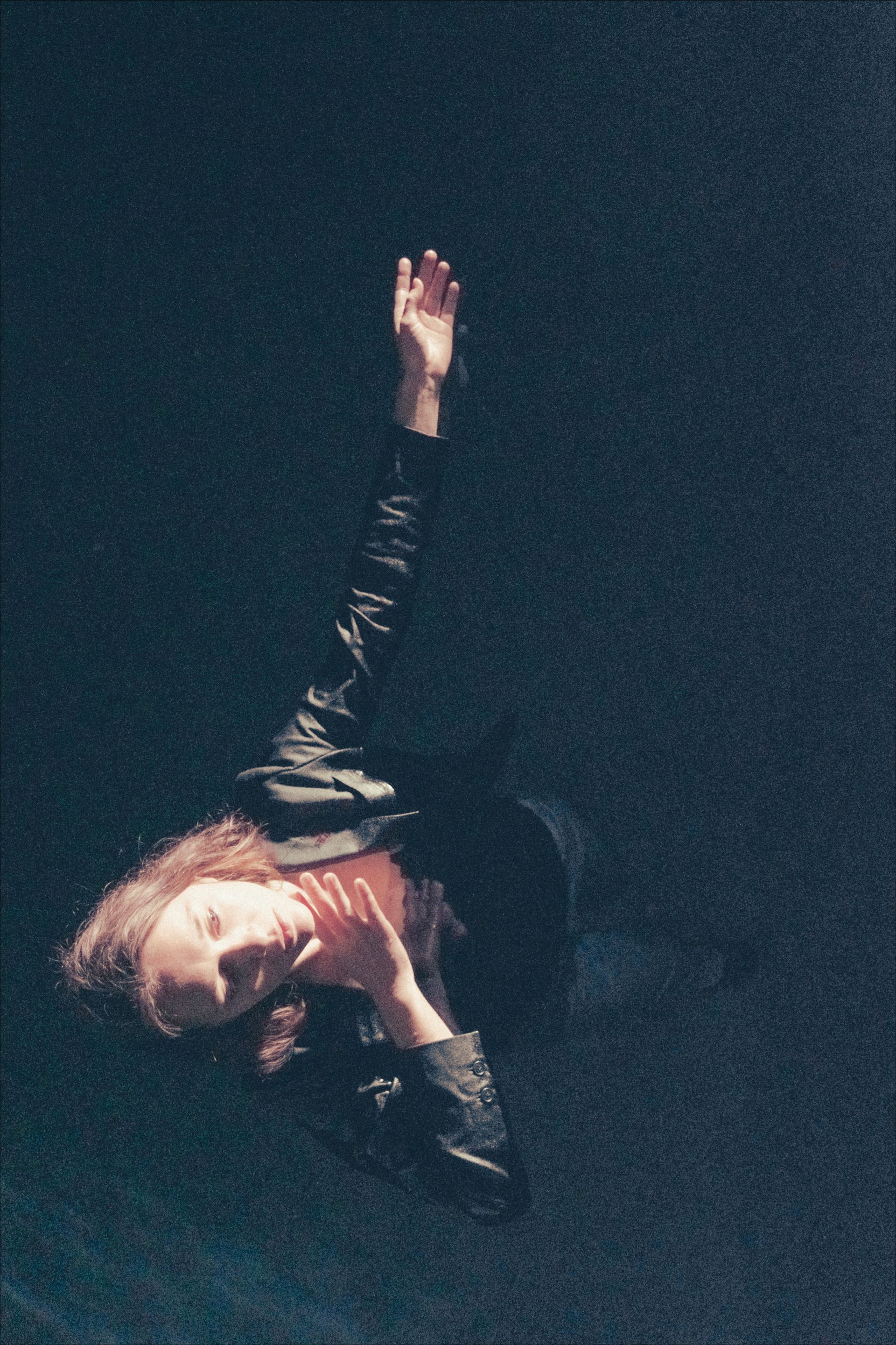 Woman reaching upwards, lit from above, in dark setting.