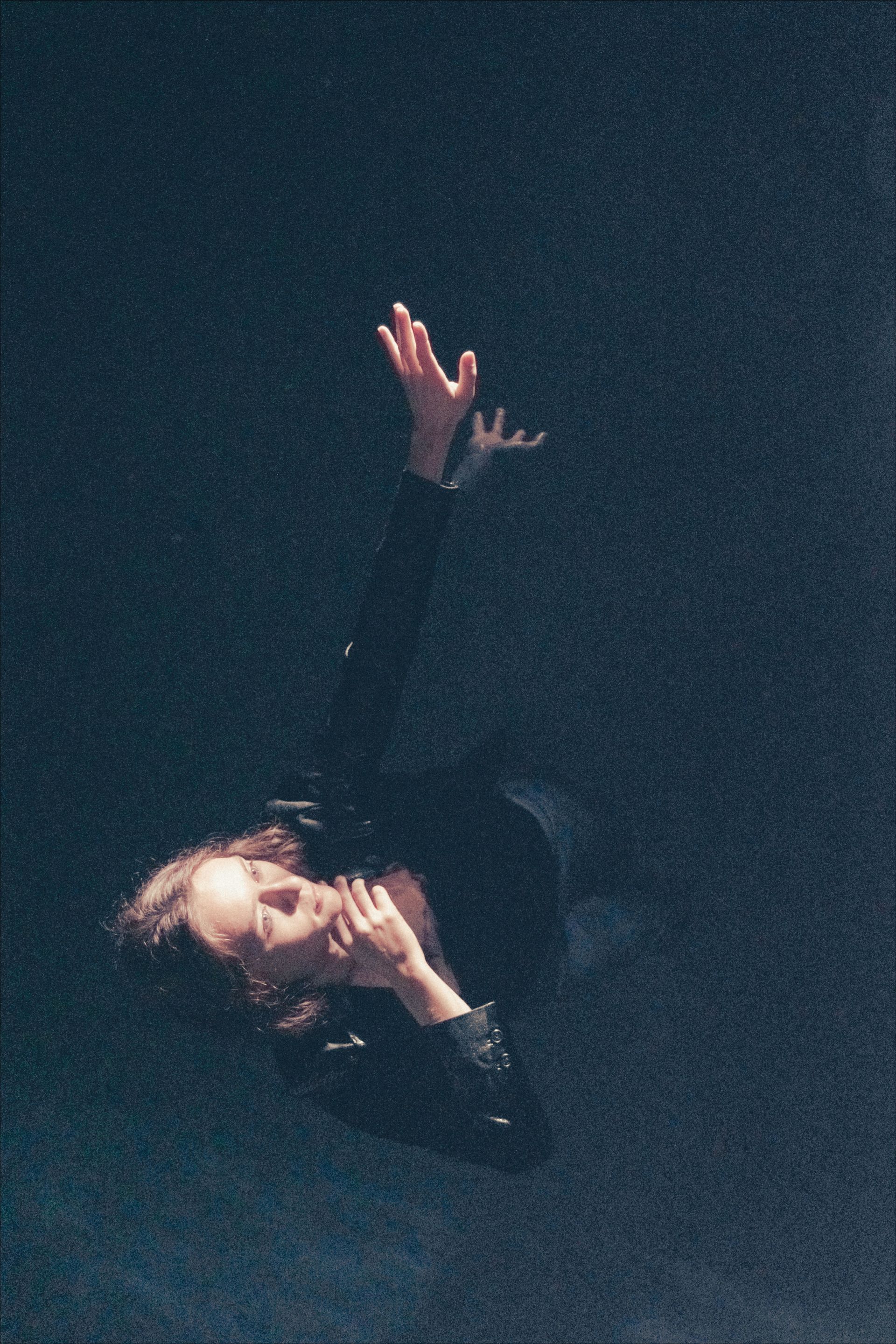 Person with arms raised, in dark water or space. Eyes closed, looking up.