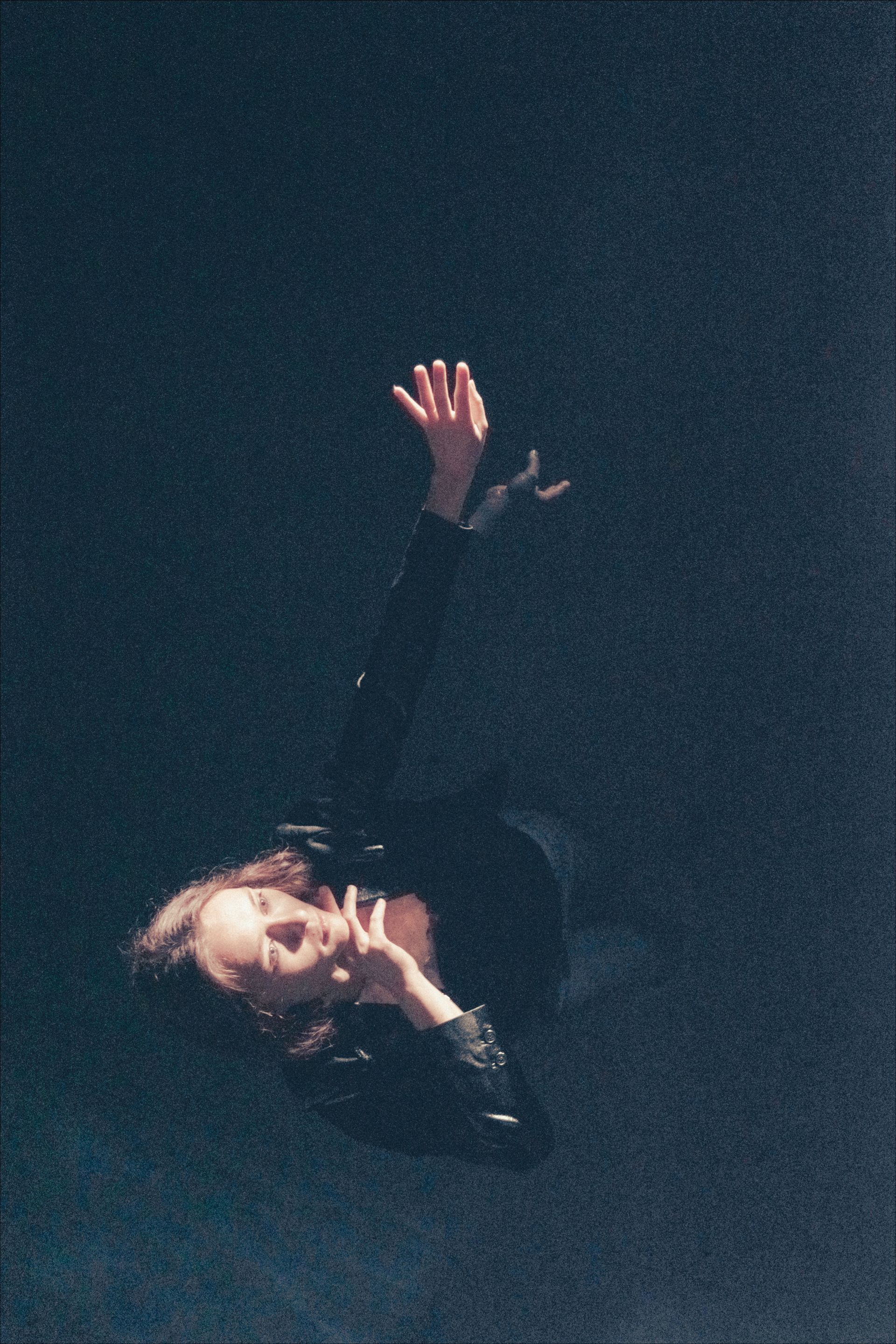 Person in dark clothing, arms raised, floating in a dark space, slight smile, head tilted upward.