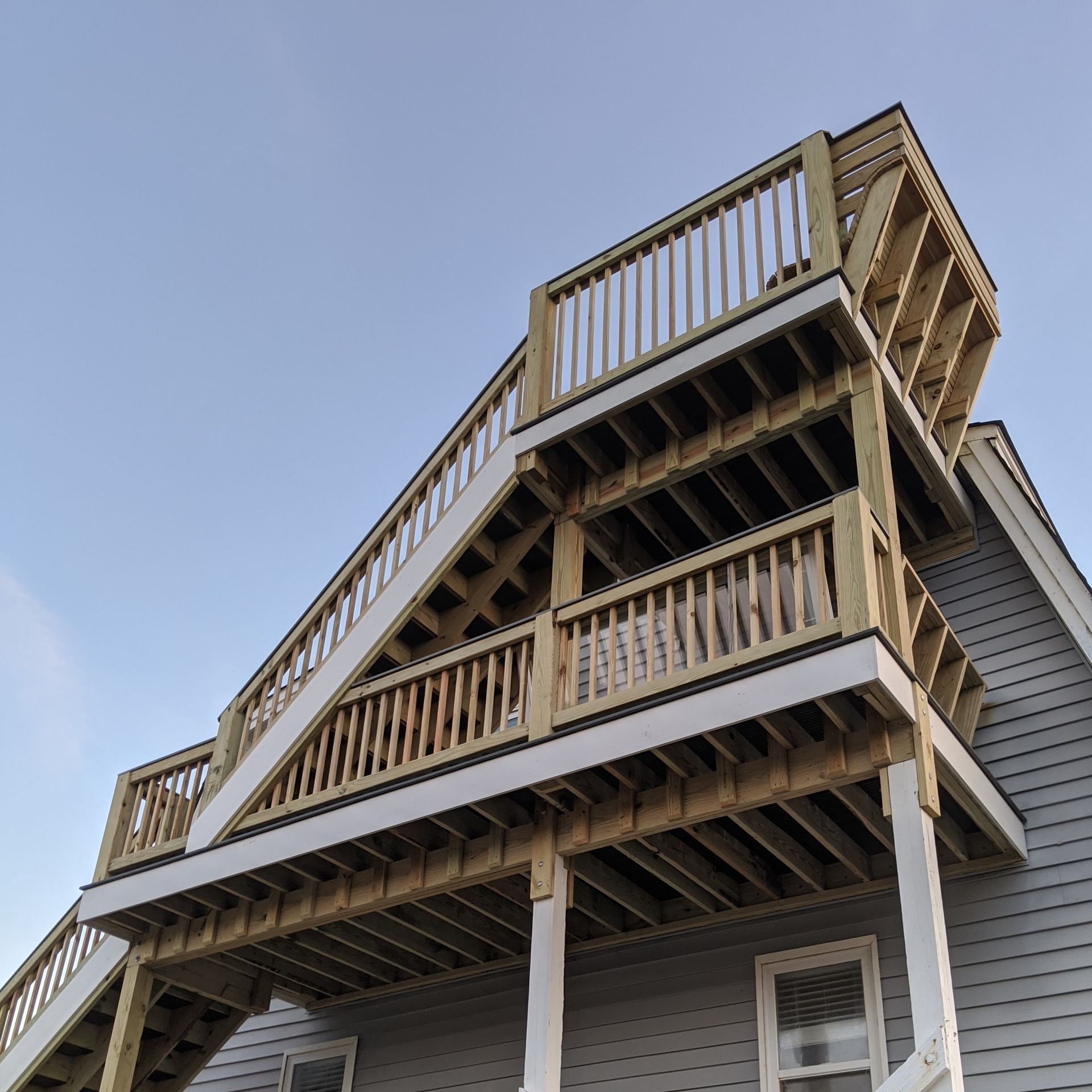 Multi-level wooden deck and stairs attached to a house with gray siding. Light blue sky in the background.
