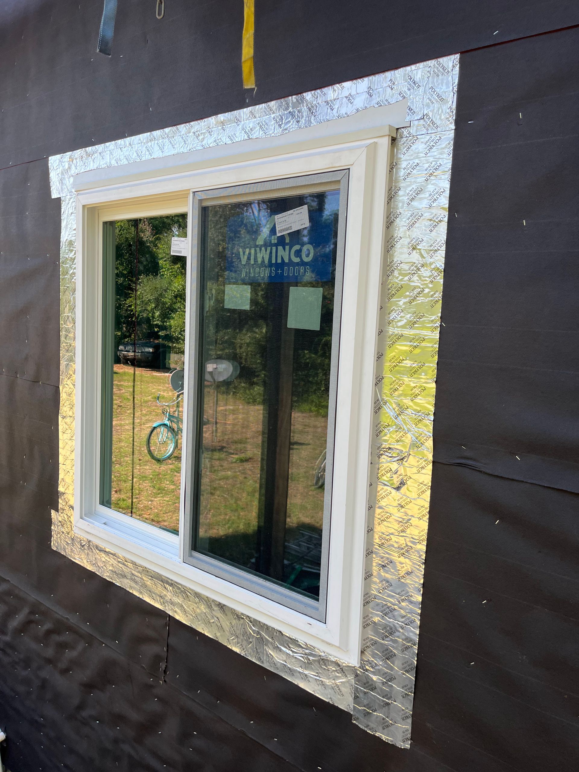A white sliding window installed in a dark-walled building, wrapped with silver flashing tape.