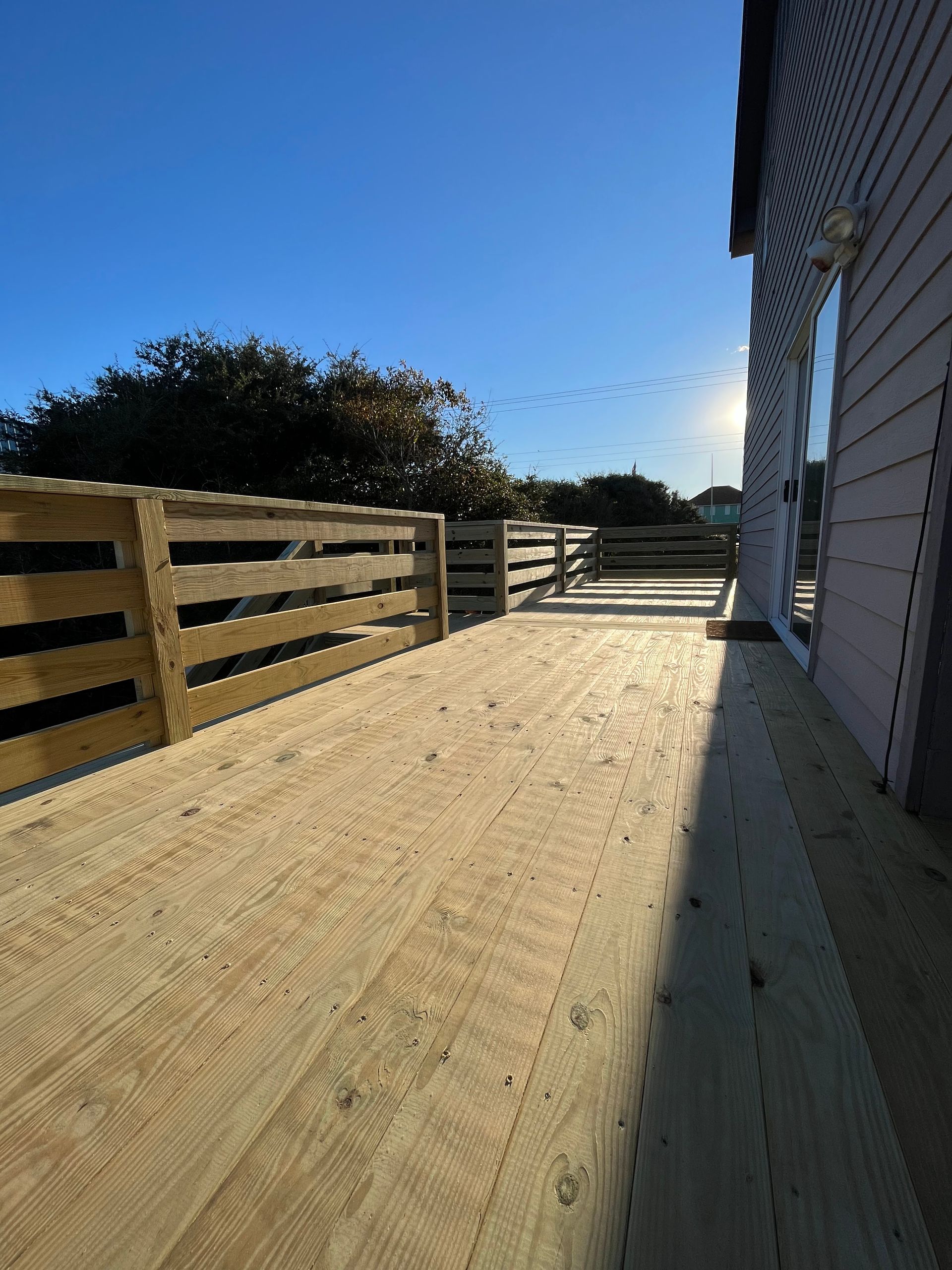 Wooden deck with a fence on one side, next to a light-colored house, under a clear blue sky.